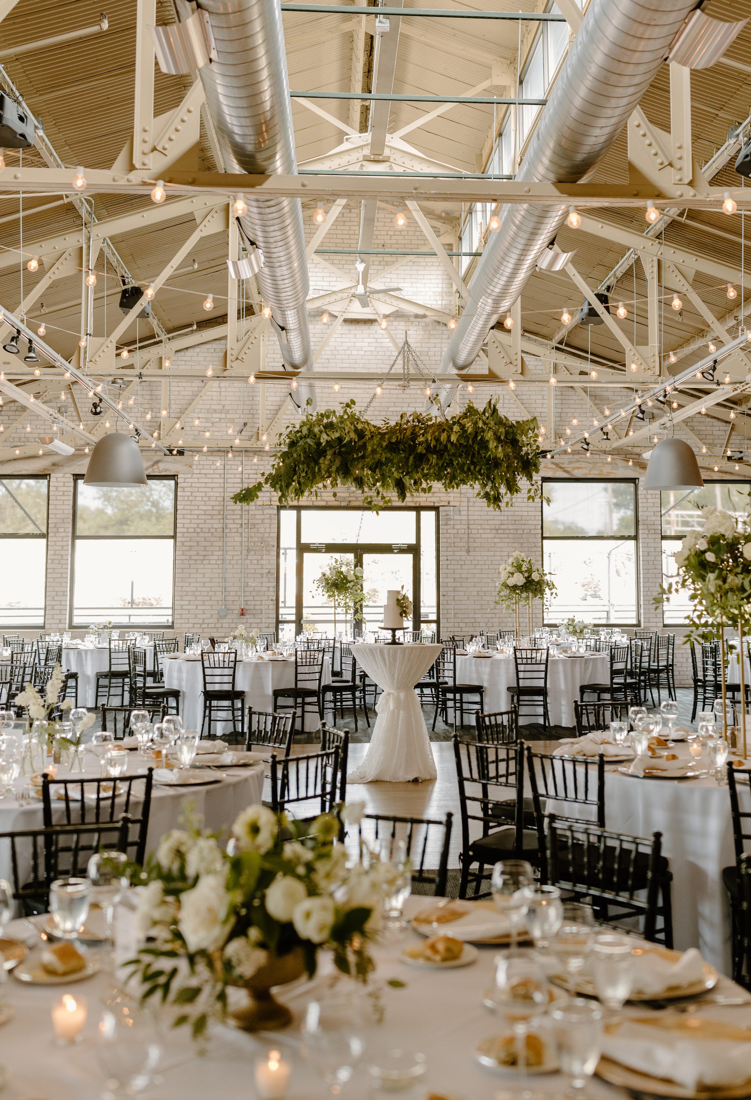 Wedding reception setup with round tables and white tablecloths, floral centerpieces, and black chairs in a spacious industrial-style venue with exposed ductwork and string lights.