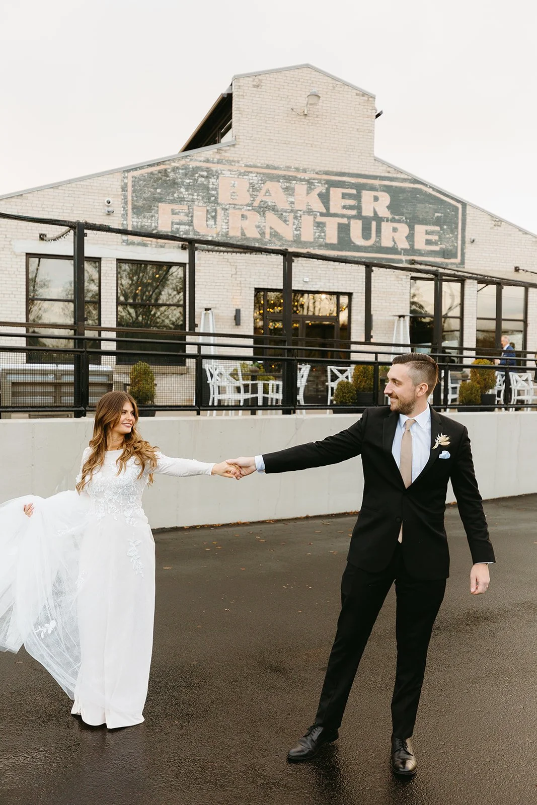 A bride and groom holding hands in front of a building with a sign that reads 'Baker Furniture.' The bride is in a white wedding dress, and the groom is in a black suit. They are smiling and looking at each other.
