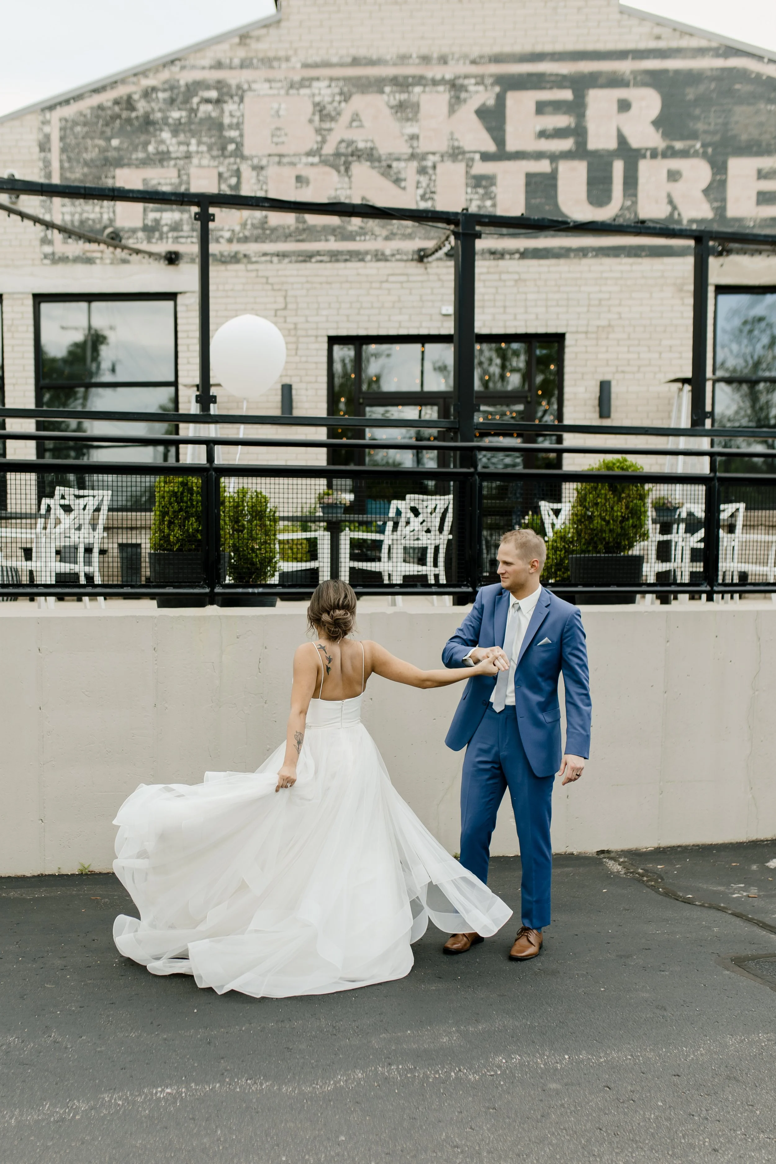 A bride and groom dancing outdoors in front of a beige wall and a building with a brick wall. The bride wears a white wedding gown with a full skirt and has tattoos on her back and arm. The groom wears a blue suit and brown shoes. They hold hands whi