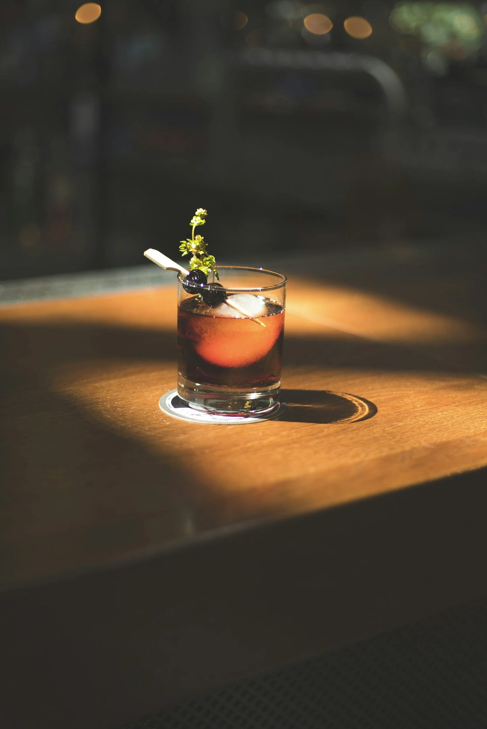 A glass of dark red cocktail garnished with a sprig of herbs and blueberries, sitting on a wooden surface with warm lighting and a blurred background.