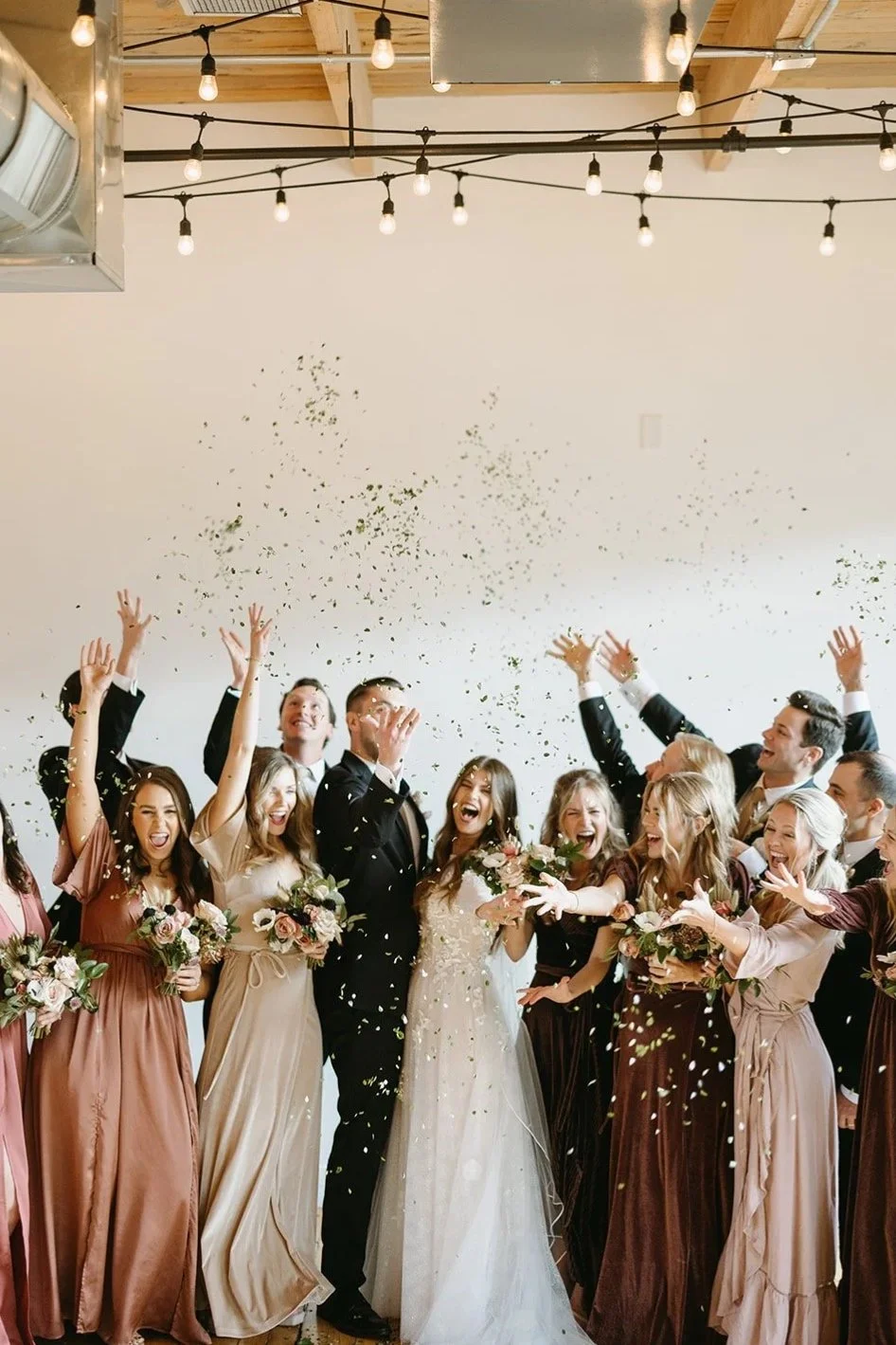 Group of wedding guests celebrating with confetti in a room with hanging string lights and wooden ceiling beams.