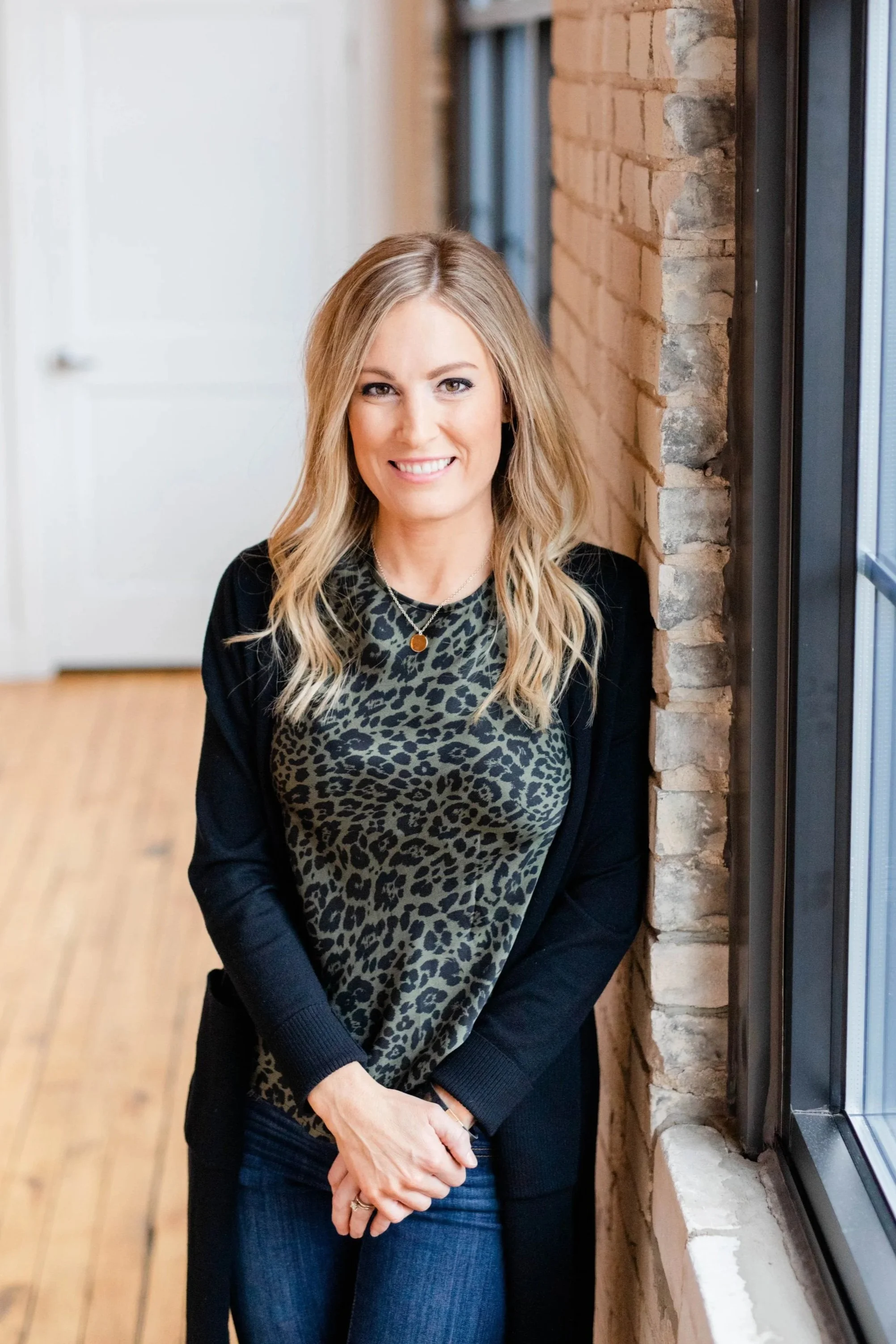 A woman with long blonde hair smiling, wearing a dark blazer and leopard print top, standing near a brick wall and window inside a building.
