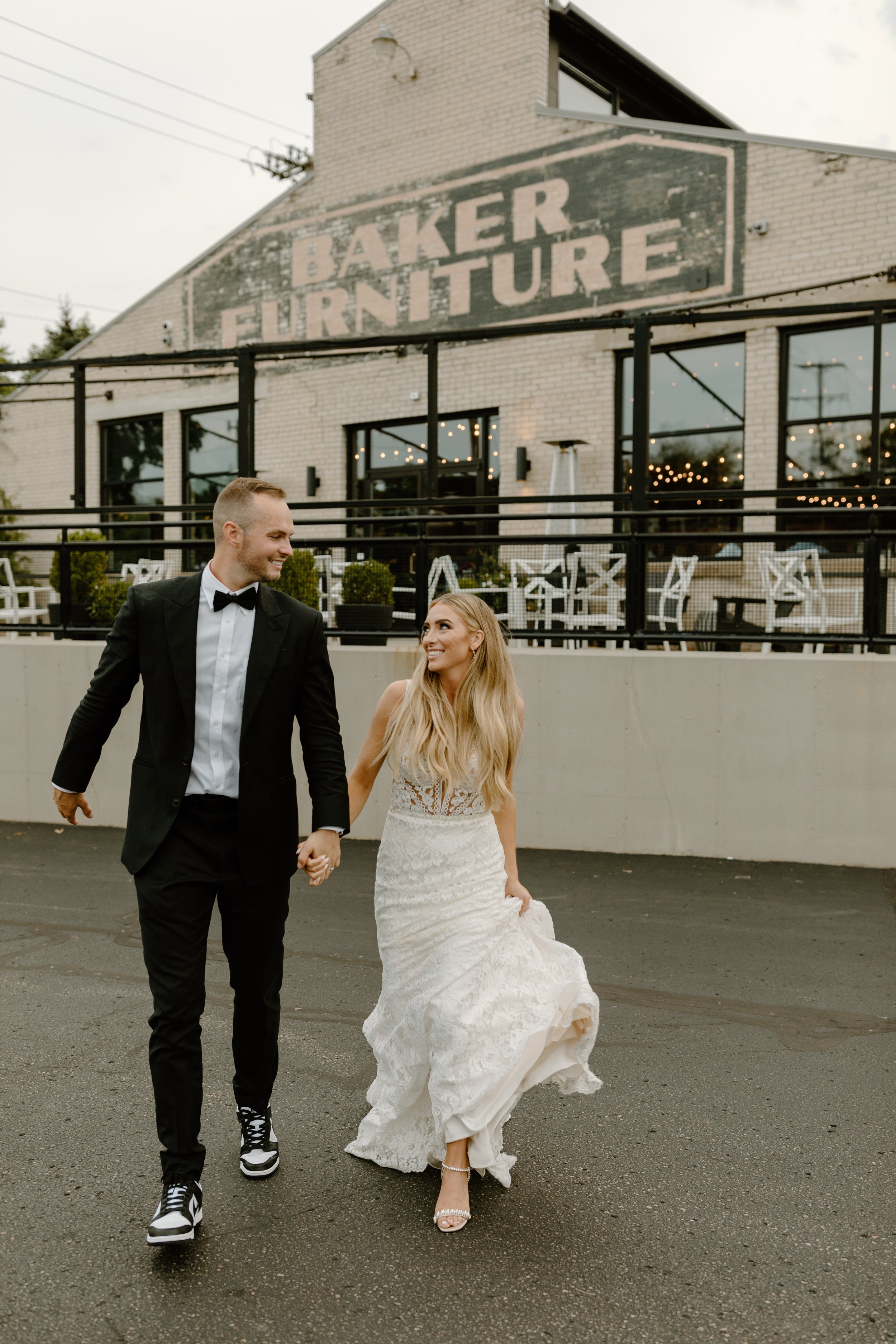 A newlywed couple walking hand in hand outside a furniture store called 'Baker Furniture.' The groom is wearing a black tuxedo with a bow tie and sneakers, while the bride is wearing a white lace wedding dress with high-heeled sandals. They are smili