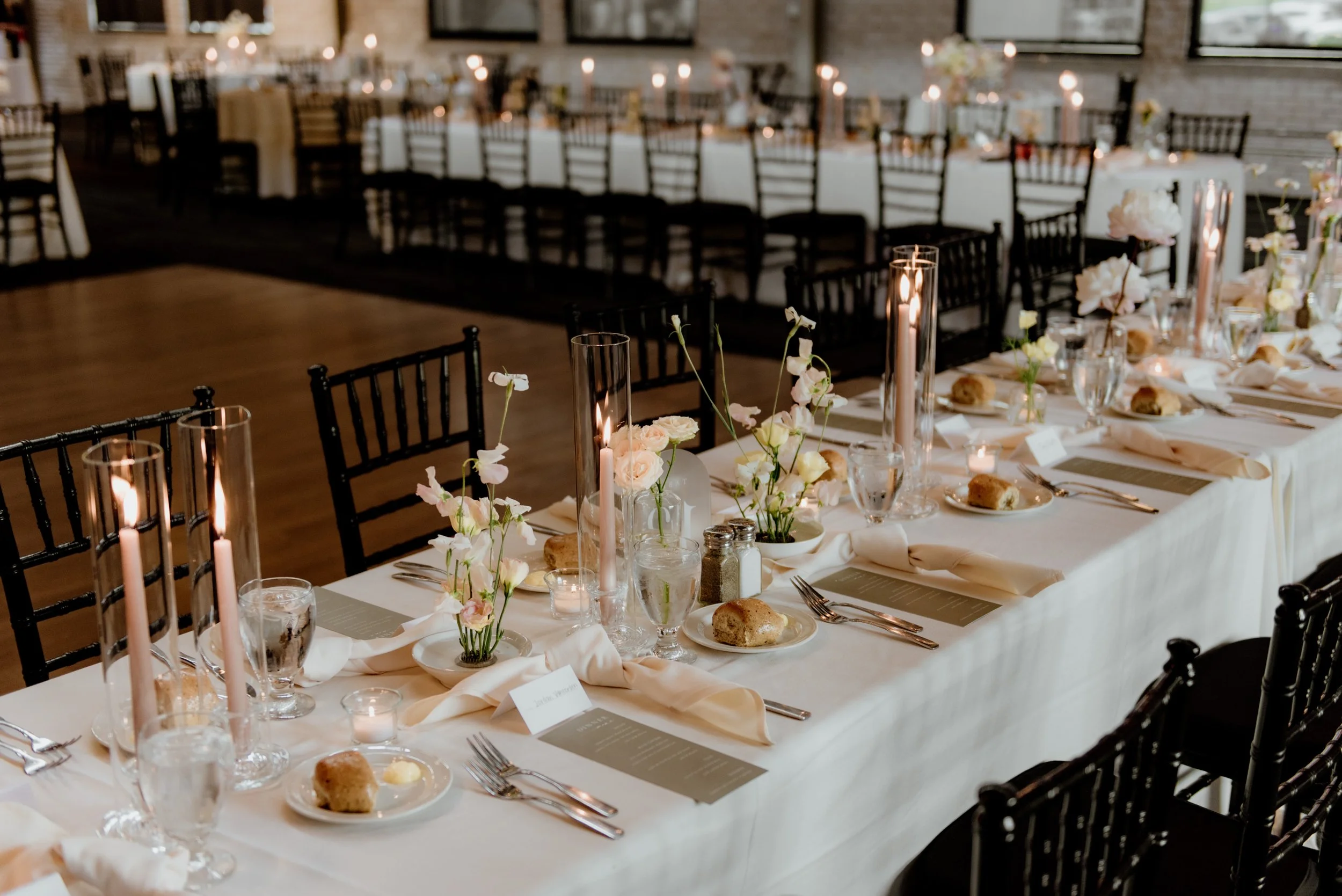 A long banquet table set for a formal event with white tablecloths, floral centerpieces, candles in tall glass holders, glassware, plates with bread rolls, and place cards. Black chairs surround the table, and in the background there are more chairs 