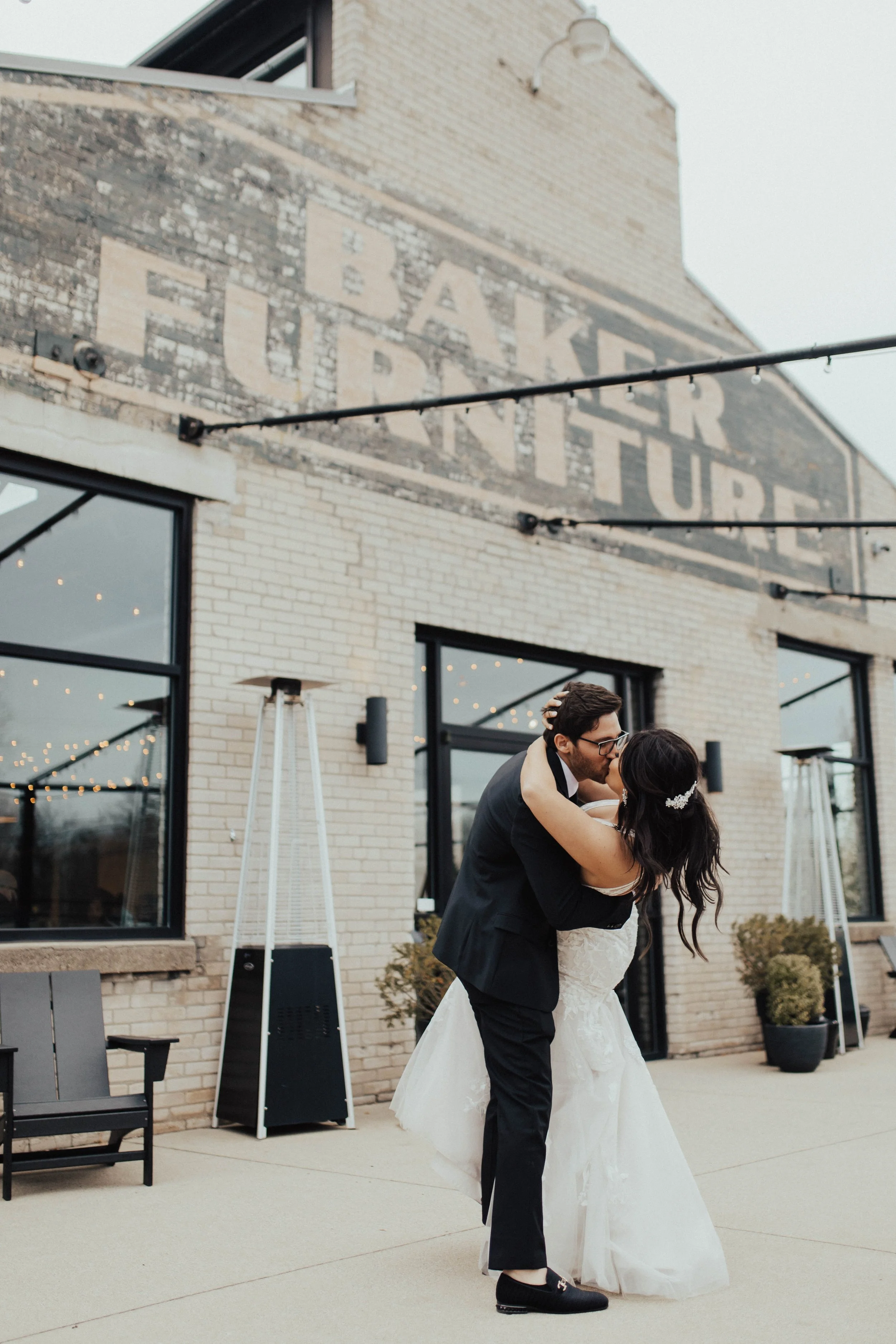 A bride and groom share a kiss outdoors in front of a brick building, with the groom dipping the bride. The building has large windows and string lights, and the bride wears a white gown with lace details while the groom wears a black suit.