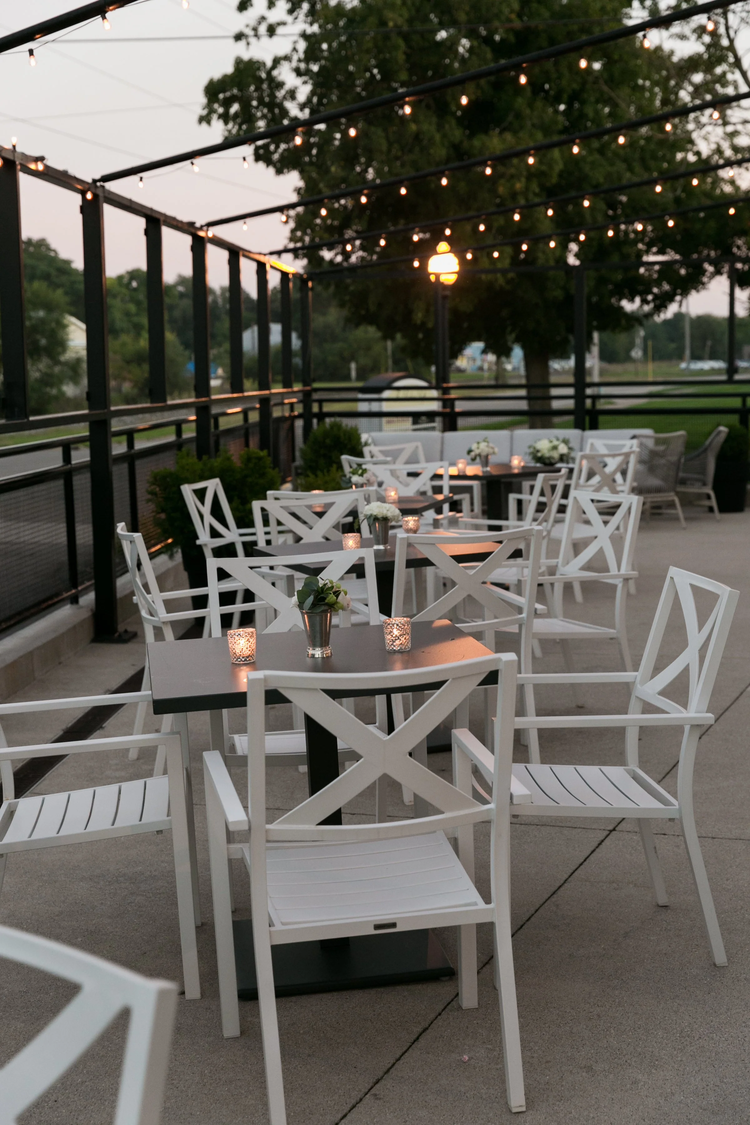 Outdoor patio with white chairs, small tables adorned with candles and floral centerpieces, string lights, and a lamppost during twilight.