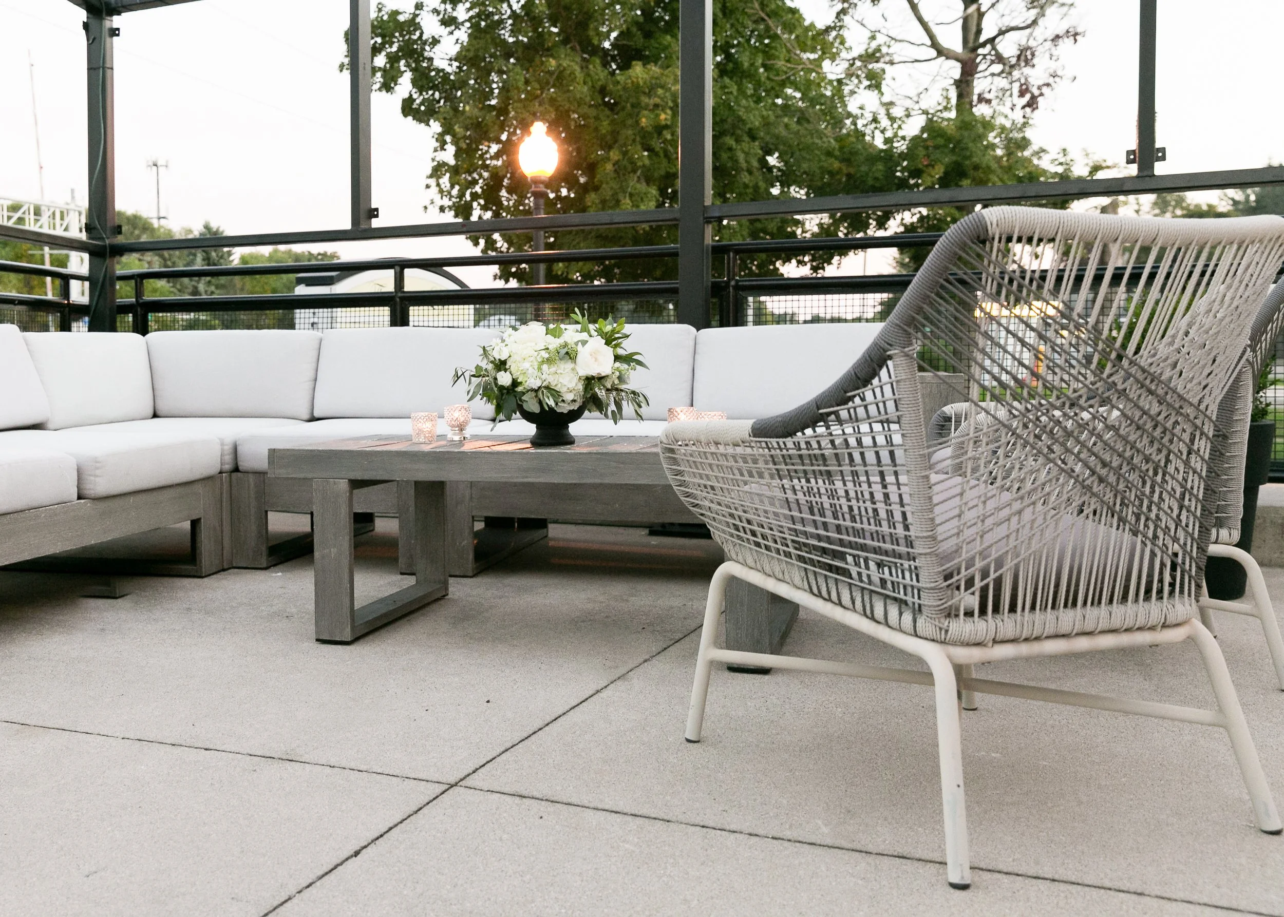 Outdoor patio furniture with a white sectional sofa, a gray woven chair, a wooden table with a large floral centerpiece, and candles, with a background of trees, a fence, and a streetlamp during sunset.