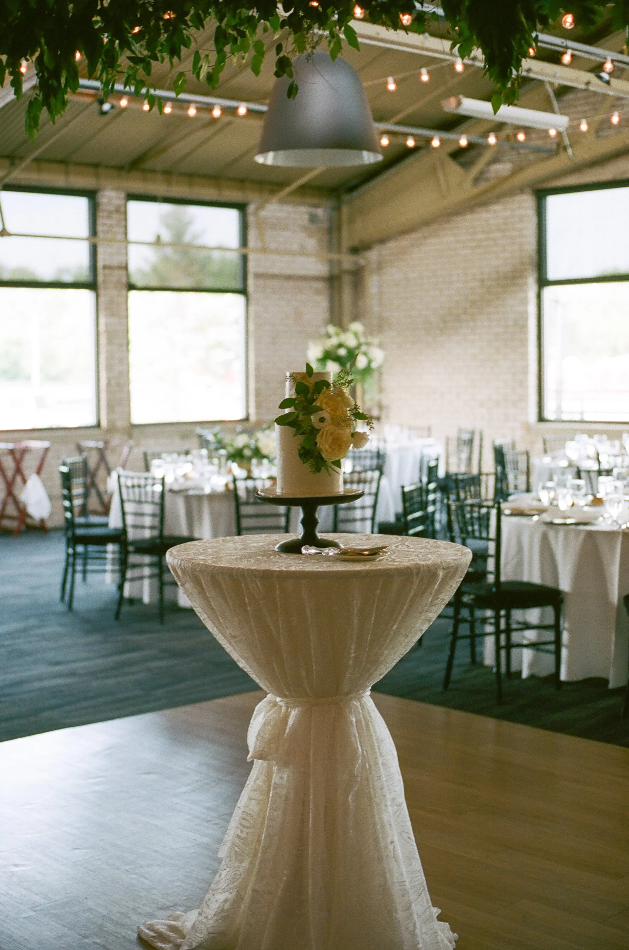 A wedding reception inside a rustic venue with large windows, where a tiered wedding cake decorated with white flowers and greenery is placed on a tall table covered with cream-colored cloth. In the background, there are tables with white tablecloths