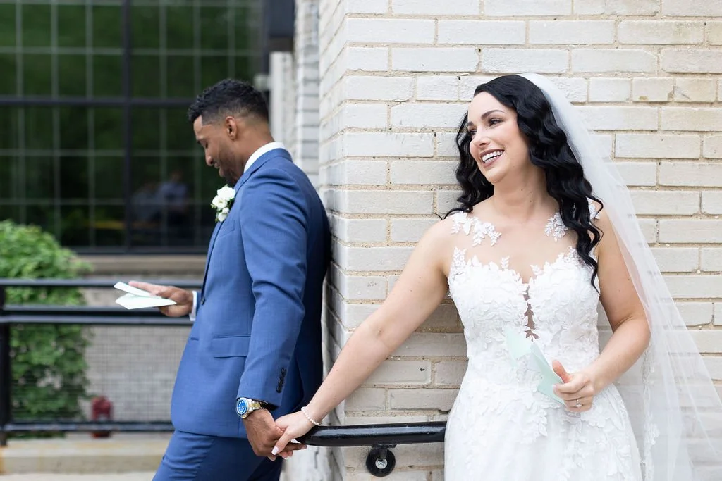 A bride and groom stand back to back outdoors near a brick wall, both holding papers, with the bride smiling and the groom looking down at his paper.