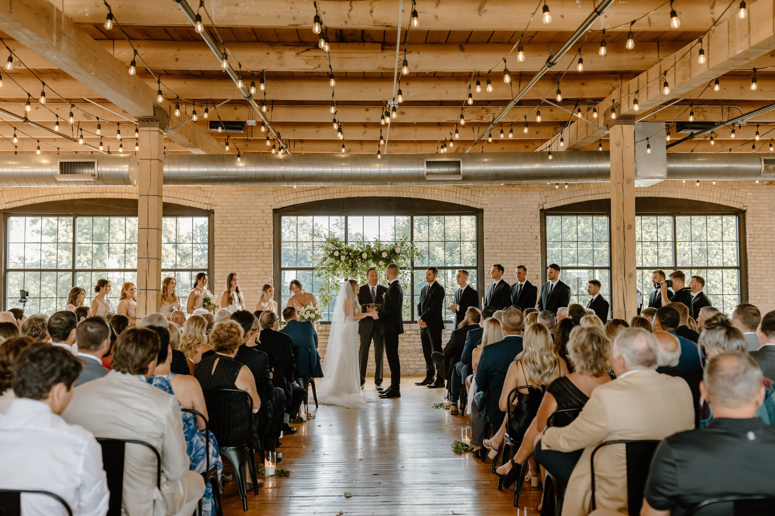 A wedding ceremony with the bride and groom exchanging vows in front of a large audience in an industrial-style venue with high windows, exposed brick walls, and wooden ceiling beams.