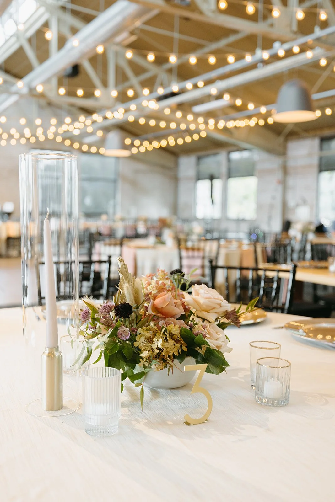Decorated table with floral centerpiece, candles, and table number, set in a rustic event space with string lights and large windows.