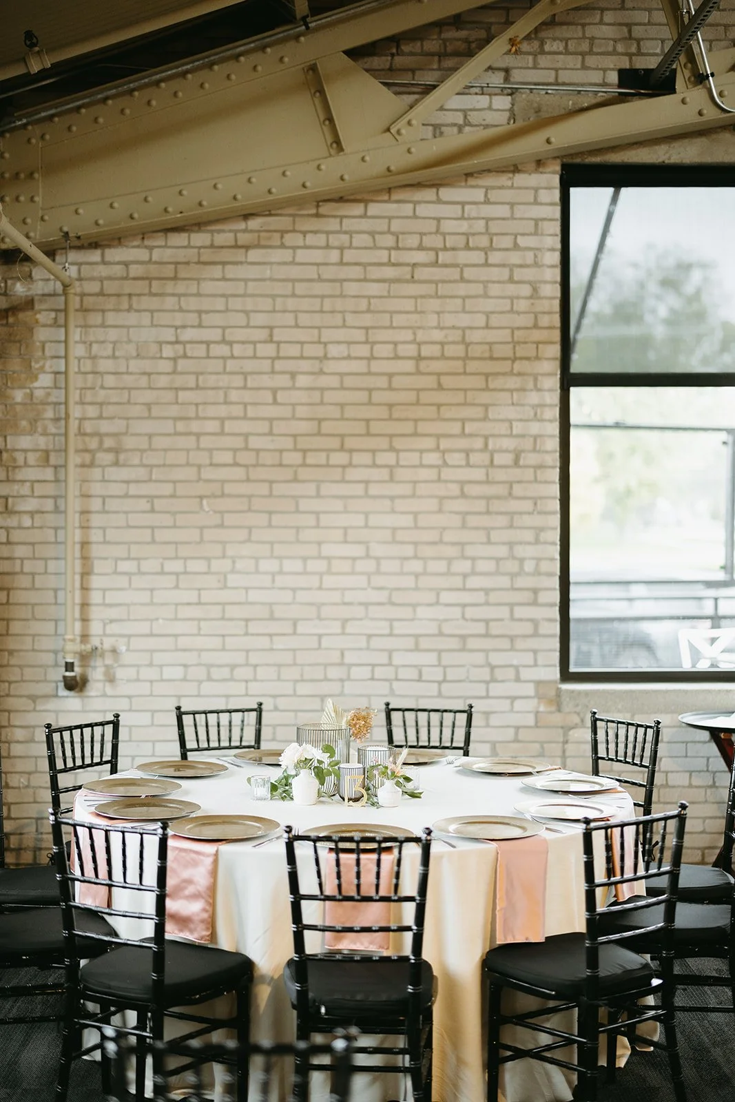 Round banquet table set for a formal event, with white tablecloths, pink and white runner, and elegant floral centerpiece, surrounded by black chairs, in a room with exposed brick walls and large windows.