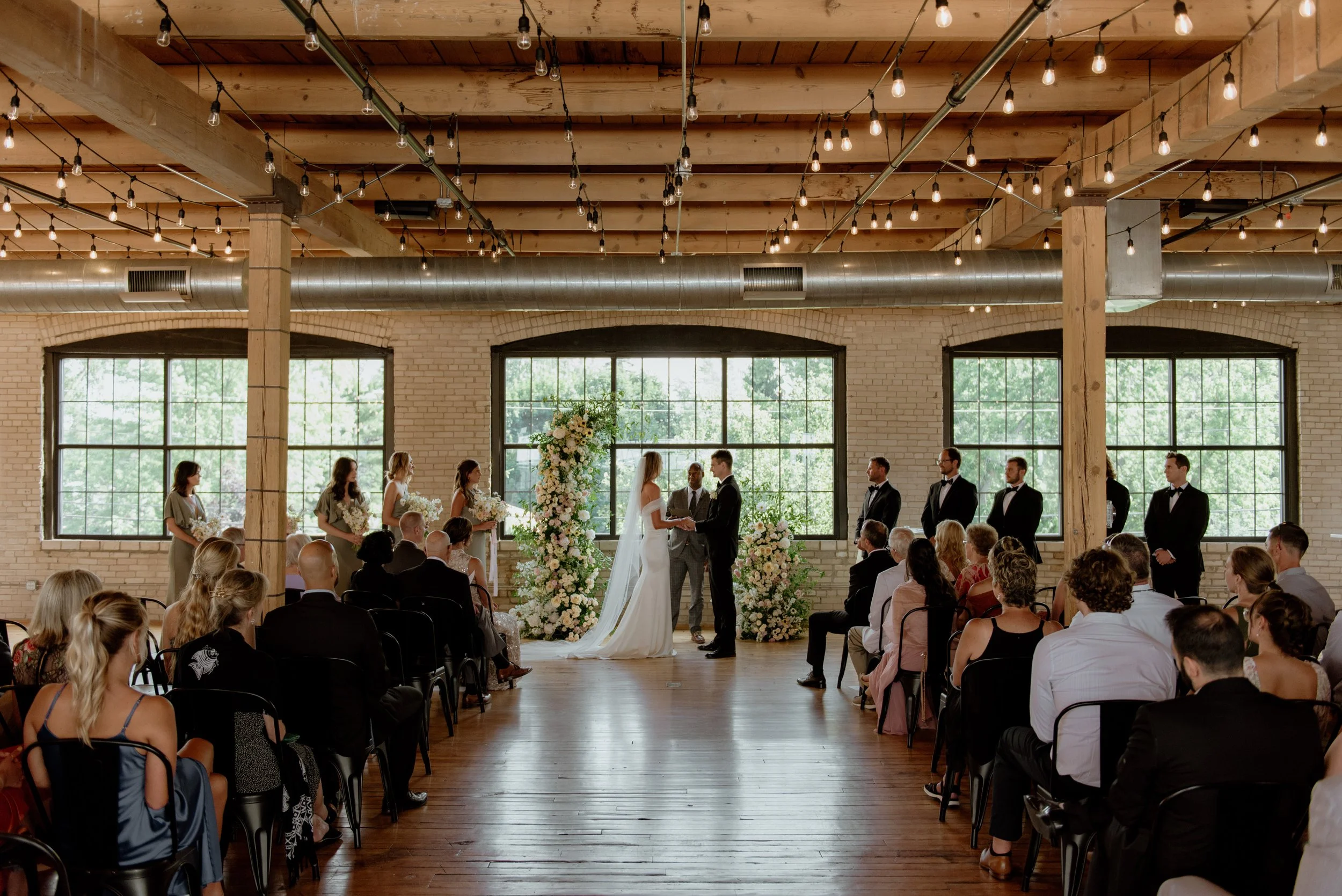 A wedding ceremony inside a spacious, industrial-style venue with exposed brick walls, large windows, wooden beams, and string lights hanging from the ceiling. The bride and groom stand at the altar, surrounded by bridesmaids and groomsmen, with gues