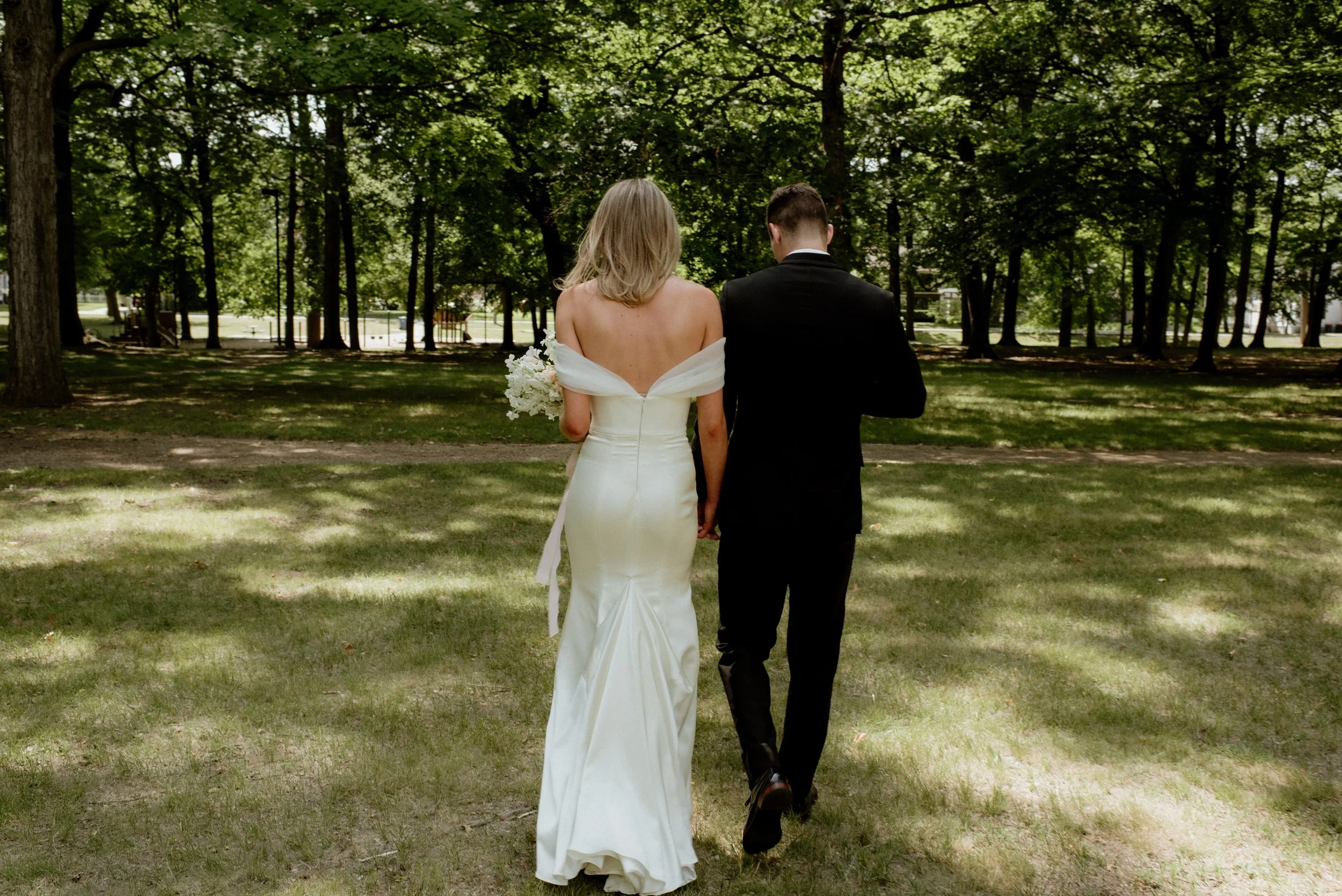 A bride and groom walking hand-in-hand through a grassy park with tall trees, seen from behind.
