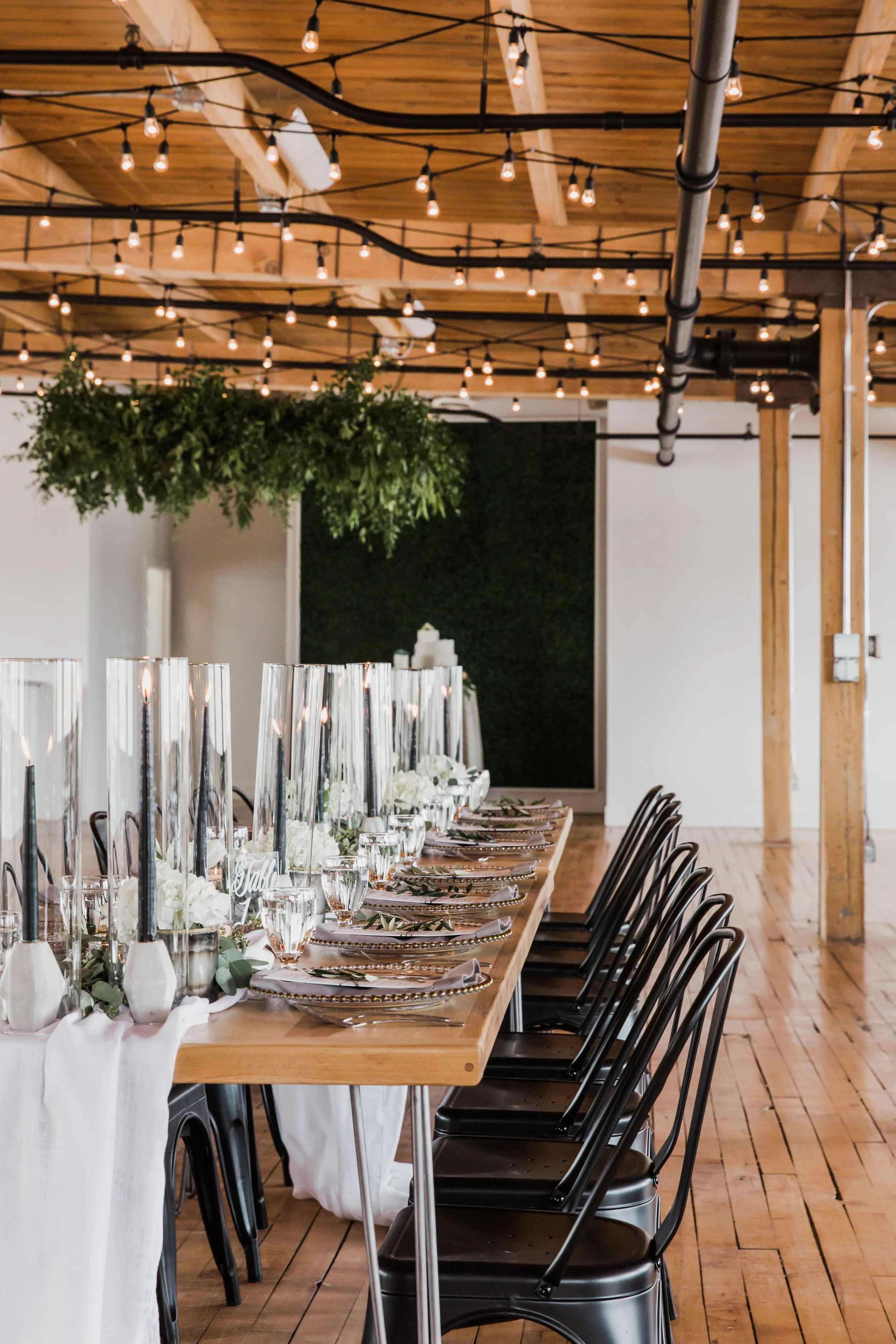 Long dining table set with white flowers, tall black candles, and glassware, in a rustic indoor venue with wooden beams, exposed piping, and string lights.