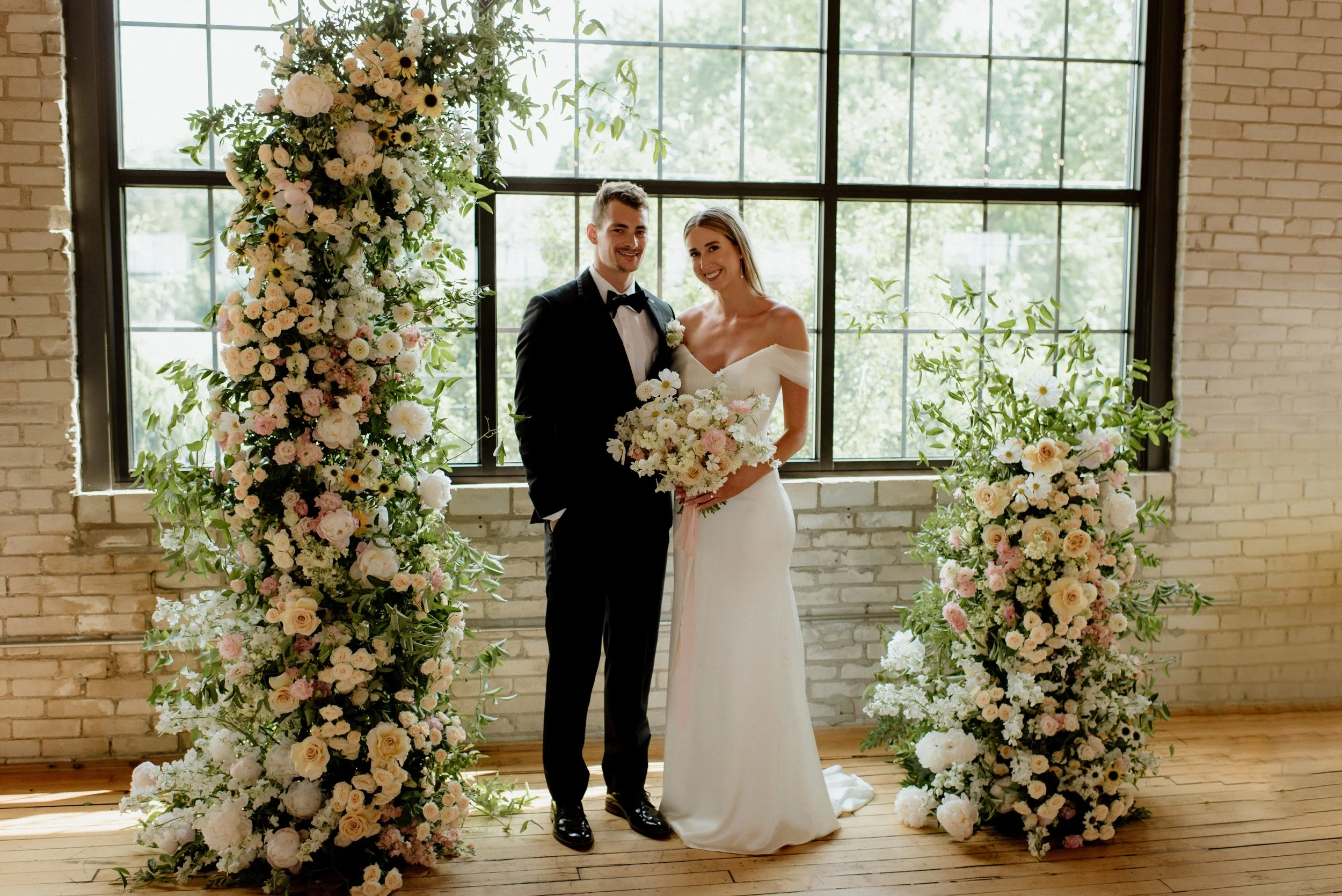 A bride and groom standing together in front of a large floral arrangement with white, blush, and yellow flowers, inside a venue with large windows and brick walls.