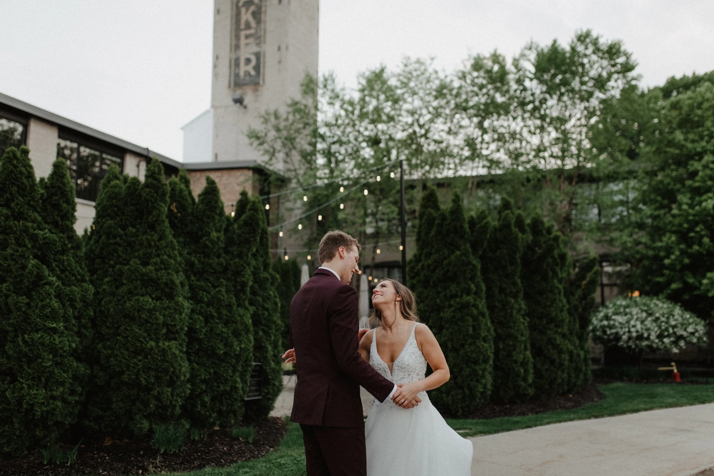 A bride and groom are outdoors, holding hands and looking into each other's eyes, with tall green shrubs and a building with string lights in the background.