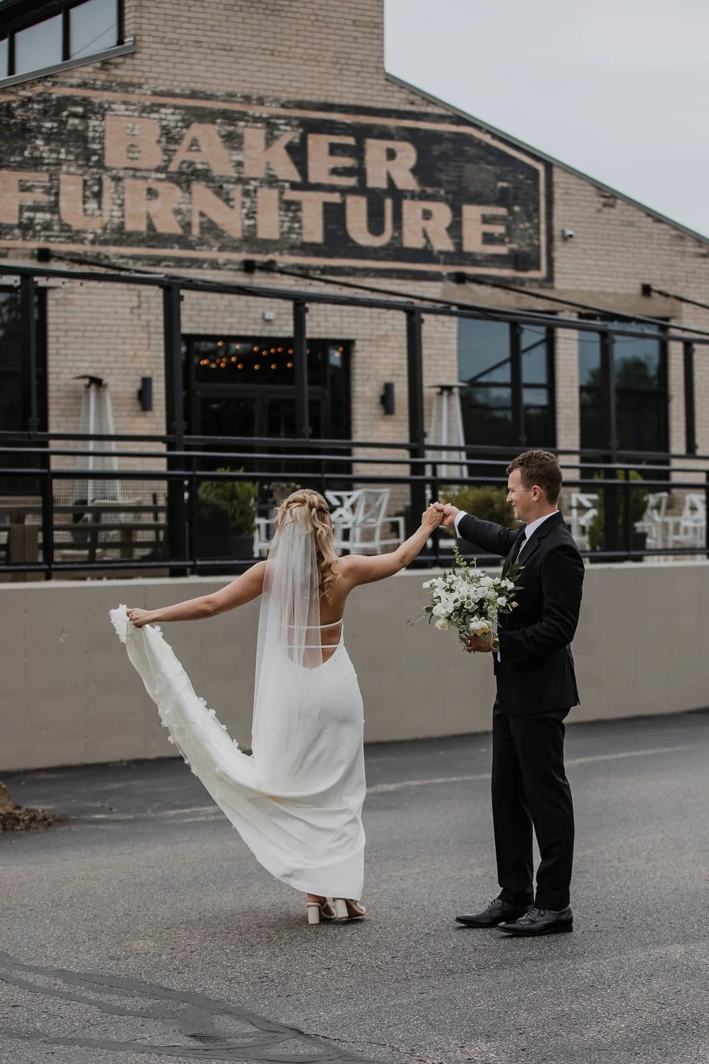 A bride and groom dancing outside in front of a brick building with a large sign reading 'Baker Furniture.' The bride is in a white wedding gown with a veil, holding her dress with one hand and extending the other towards the groom. The groom is in a