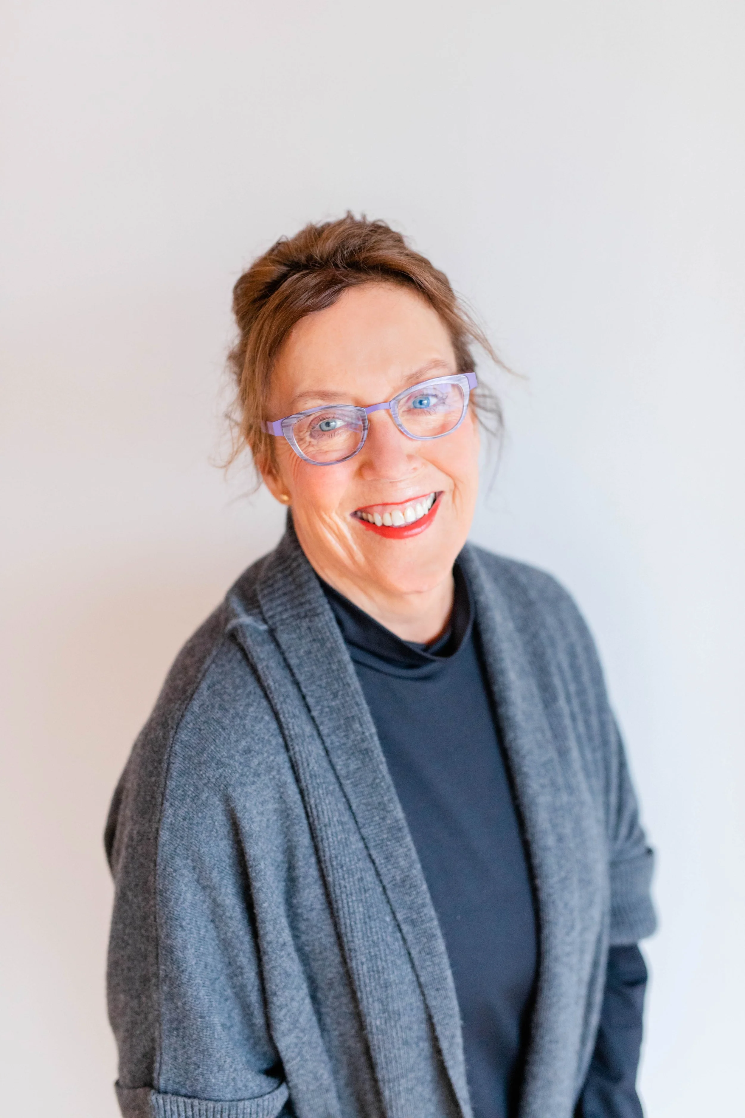 A smiling woman with short brown hair, wearing glasses with purple frames, a dark top, and a gray cardigan, standing against a plain white background.