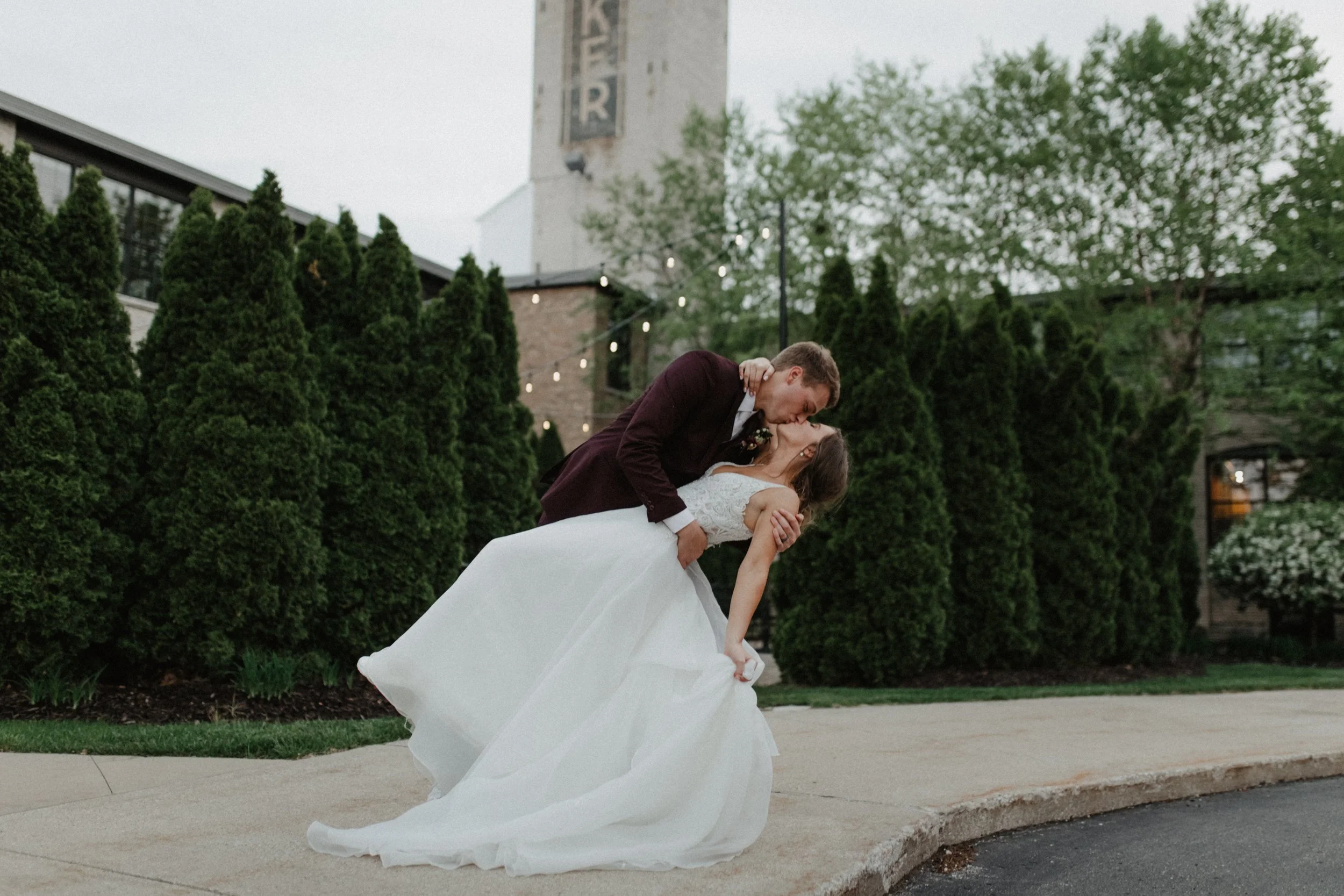 A newlywed couple sharing a kiss outdoors during their wedding, with the groom dipping the bride in front of green bushes and trees, and a building with string lights hanging in the background.