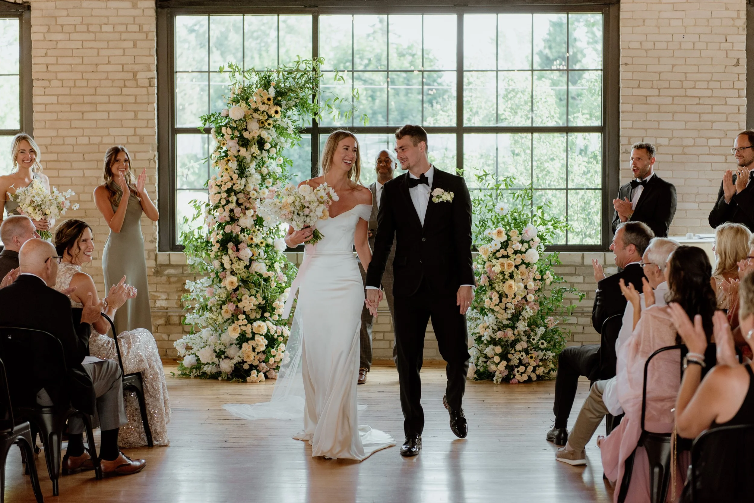 A bride and groom holding hands and smiling during their wedding ceremony in a bright, industrial-style venue with large windows and decorated with white and blush flowers.
