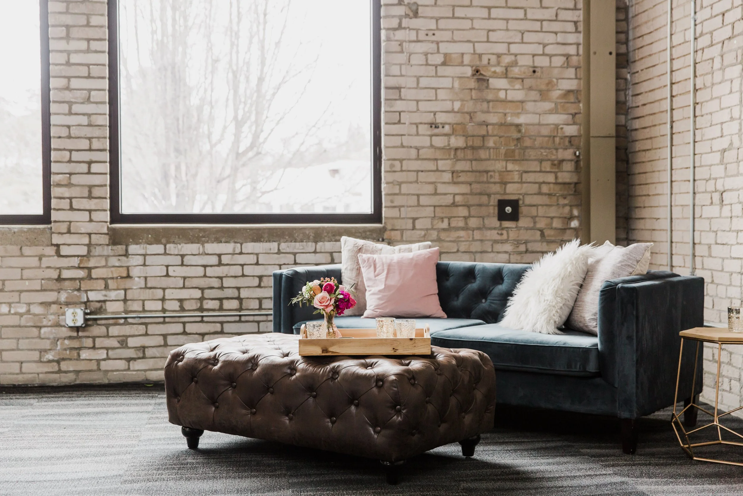 Living room with exposed brick walls, large window, dark blue velvet couch with pink and white pillows, white furry throw, wooden table with a pink floral centerpiece, and a side table with a lamp.