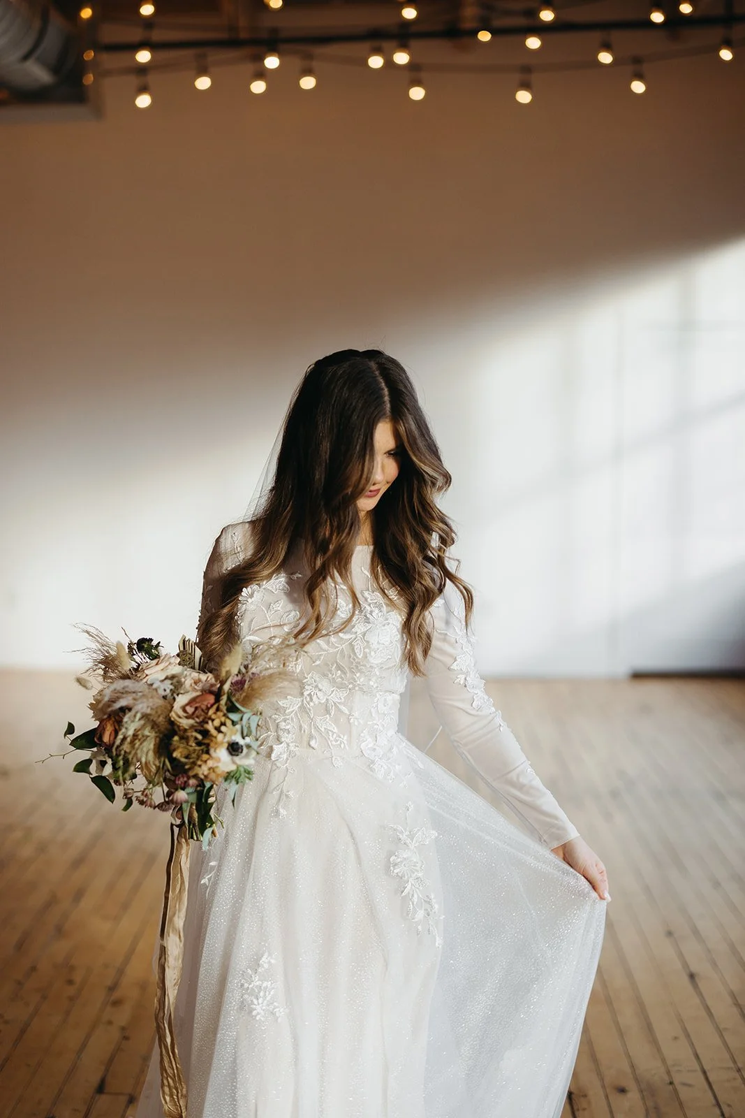 A bride in a white wedding dress with floral lace details holding a bouquet of dried flowers, standing on a wooden floor in a bright, modern space with string lights overhead.