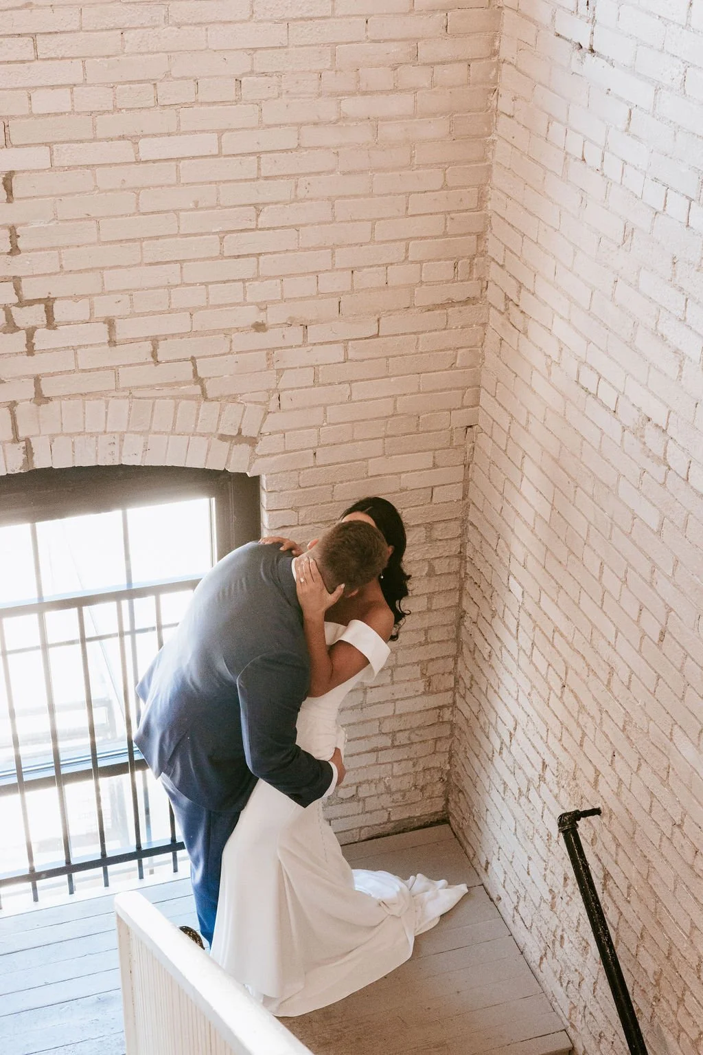 A couple is engaged in a romantic kiss in an indoor space with white brick walls and a window with metal bars, creating an intimate setting.