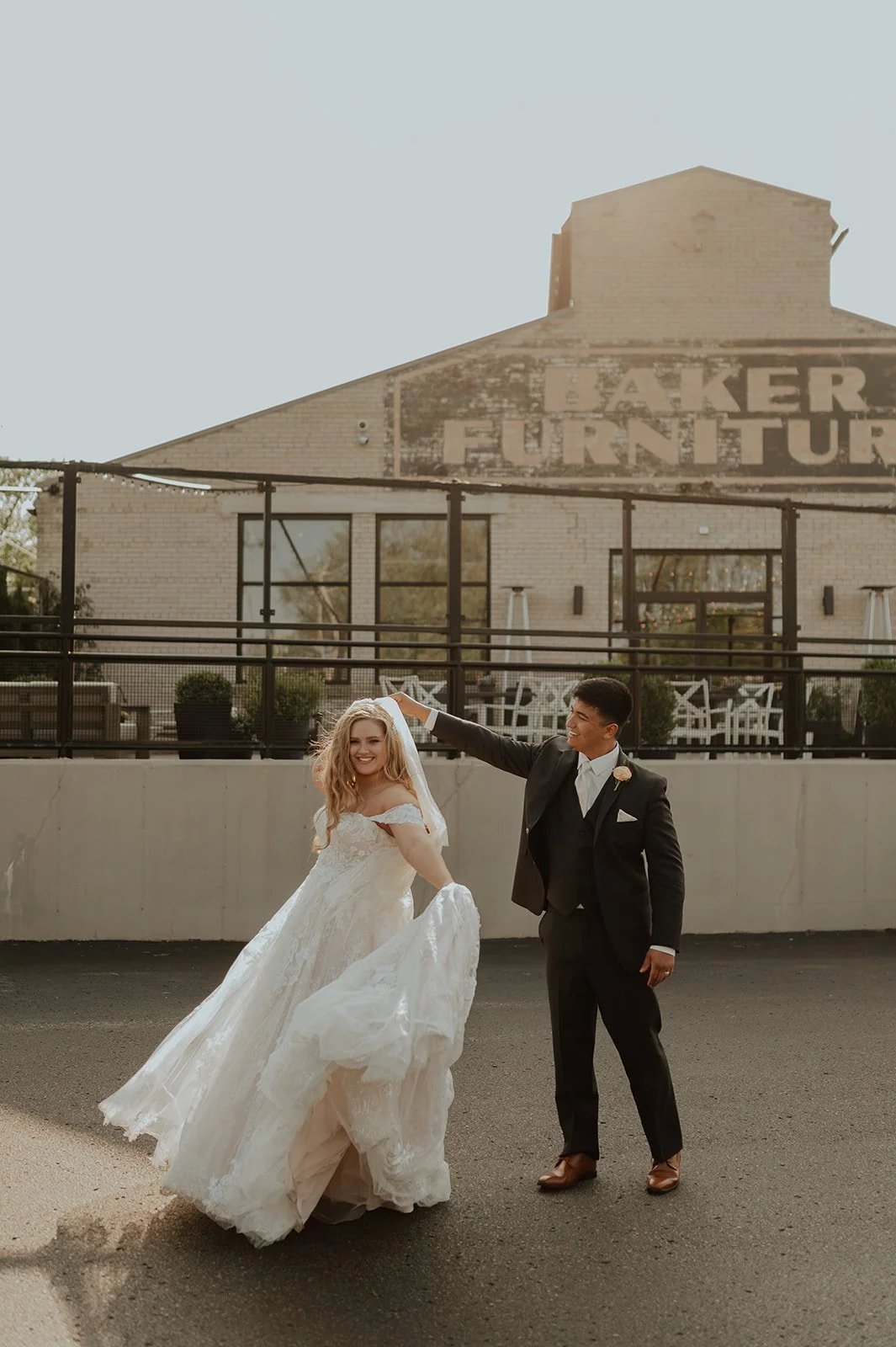A bride and groom dancing outdoors in front of a building with a sign that says 'Baker Furniture'.