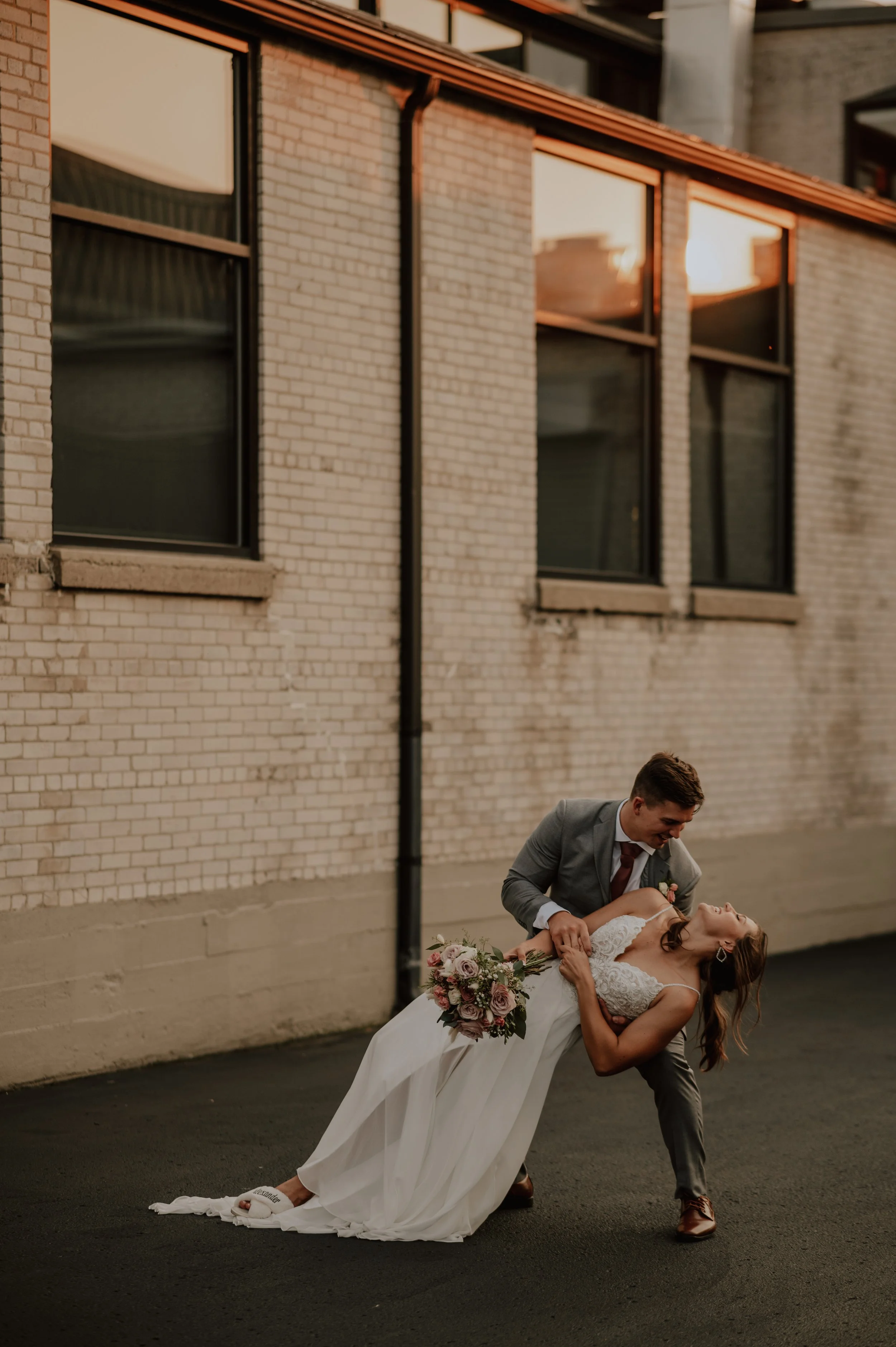 A groom dips a bride who is holding a bouquet of flowers, while they are dancing or celebrating outside against a brick building during sunset.