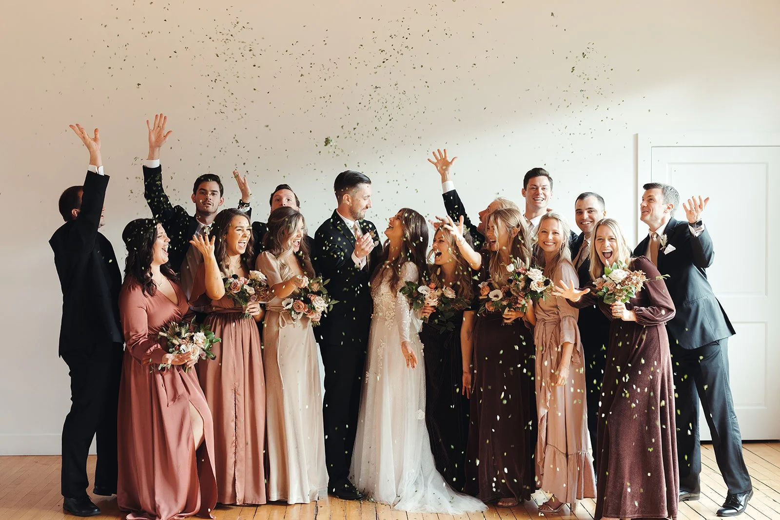Group of people celebrating a wedding, throwing confetti, smiling, and holding bouquets of flowers inside a bright room.