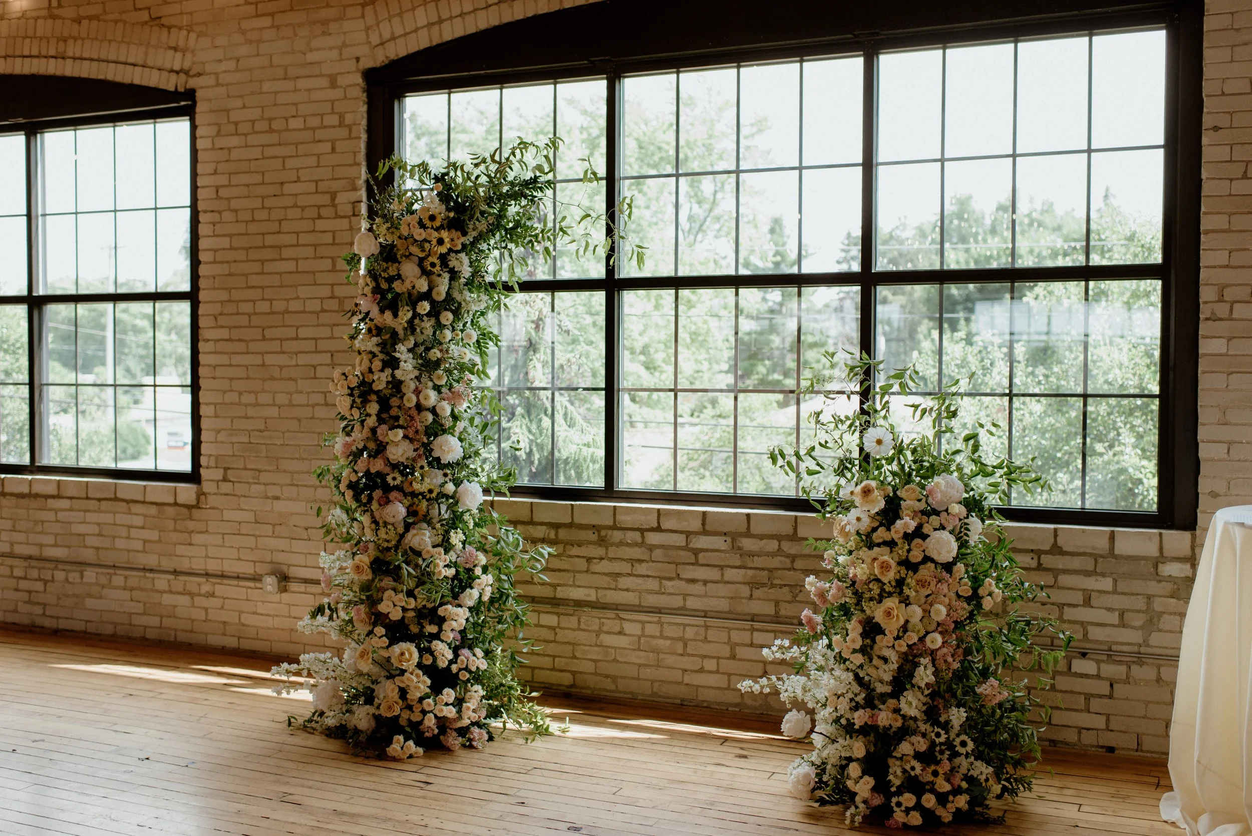 Indoor scene with large windows and white brick walls, decorated with floral arrangements of white and pale pink flowers and green foliage for a wedding or event.
