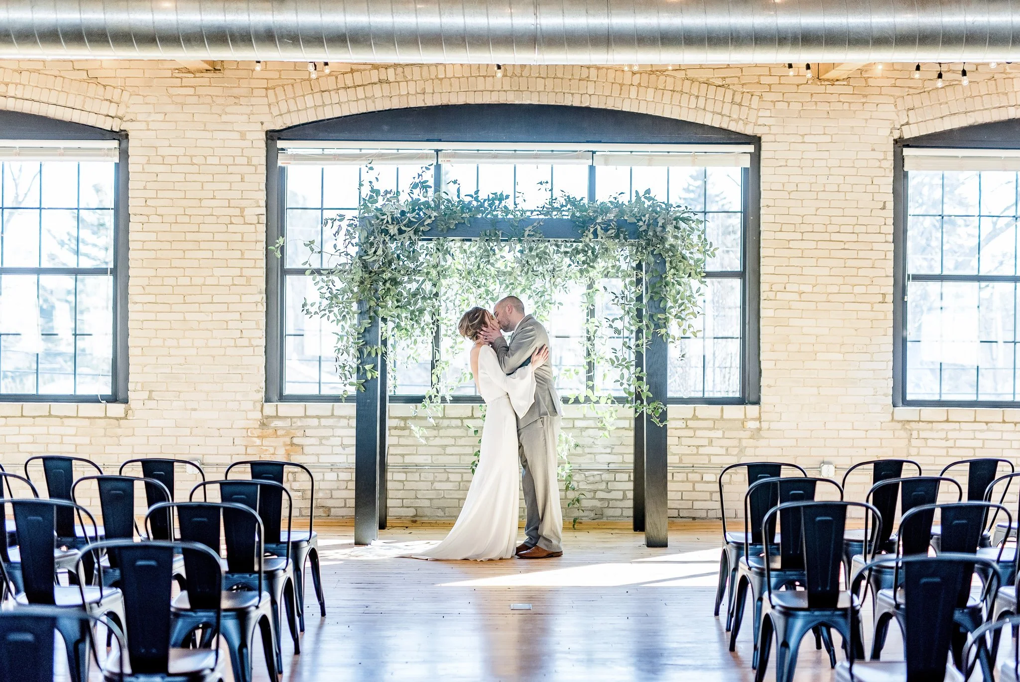 A bride and groom share a kiss at their wedding ceremony in an industrial-style venue with brick walls, large windows, and minimalist decor, including a green leafy arch behind them.