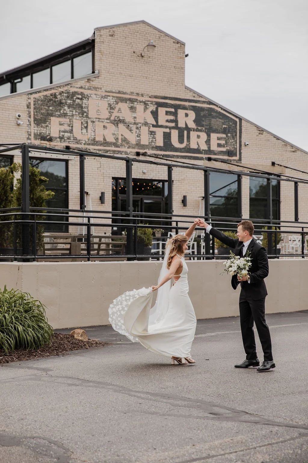 A bride and groom dancing outside a furniture store named Baker Furniture, with the groom holding a bouquet of flowers and the bride wearing a white gown, in front of a brick building with a vintage sign.