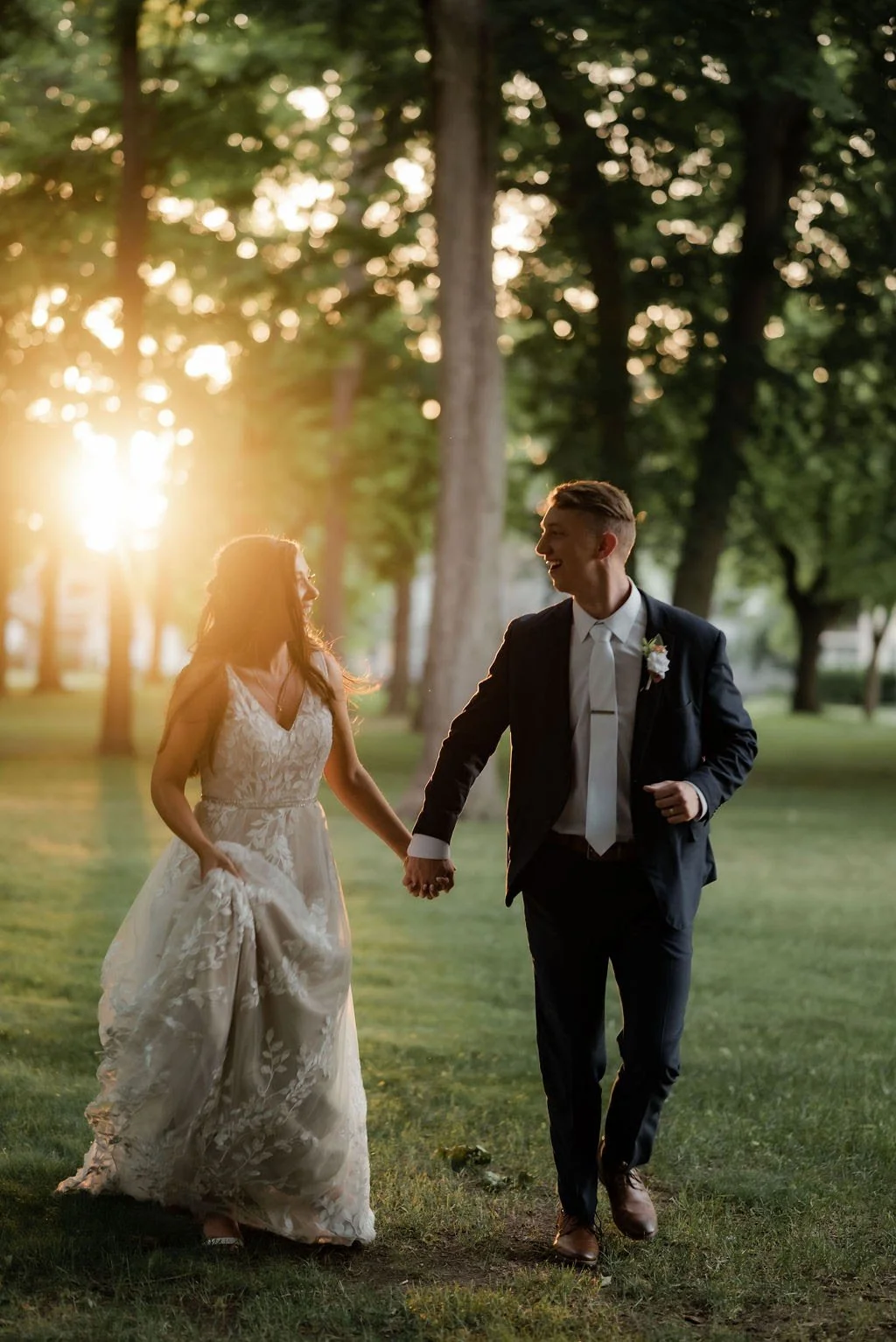 A bride and groom holding hands and walking through a park at sunset, smiling at each other after their wedding ceremony.