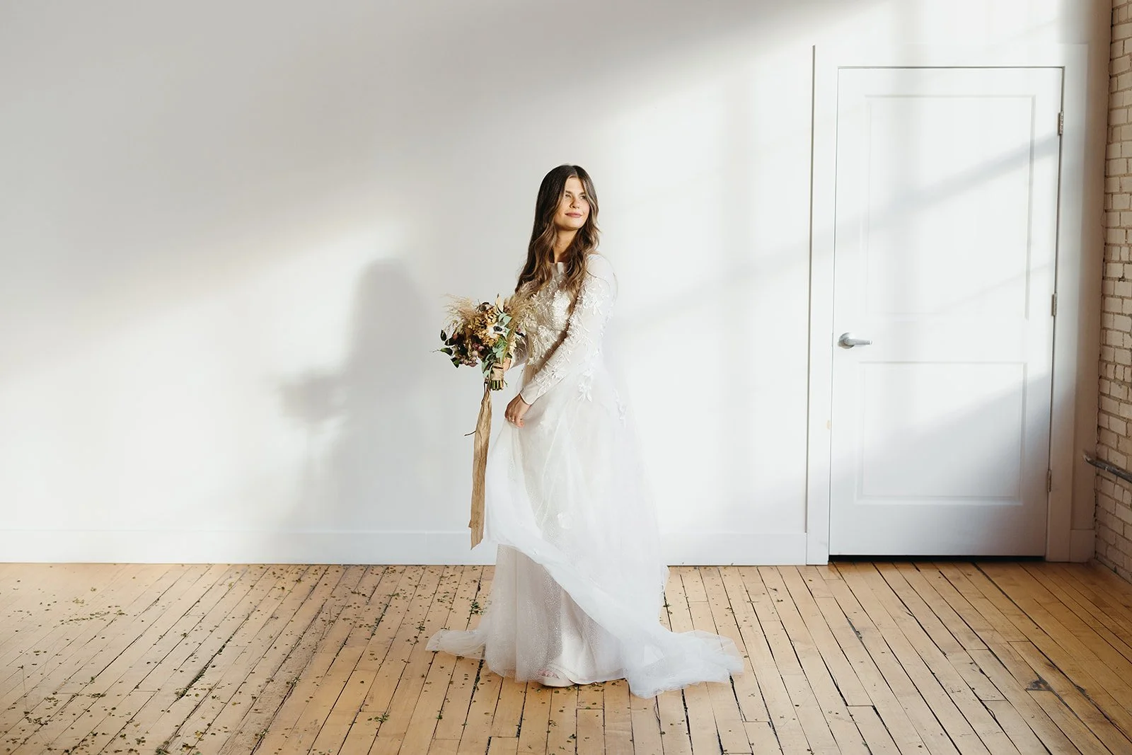 A bride in a white wedding gown holding a bouquet of flowers, standing in front of a plain white wall and a closed white door, with hardwood flooring and exposed brick wall on the right.