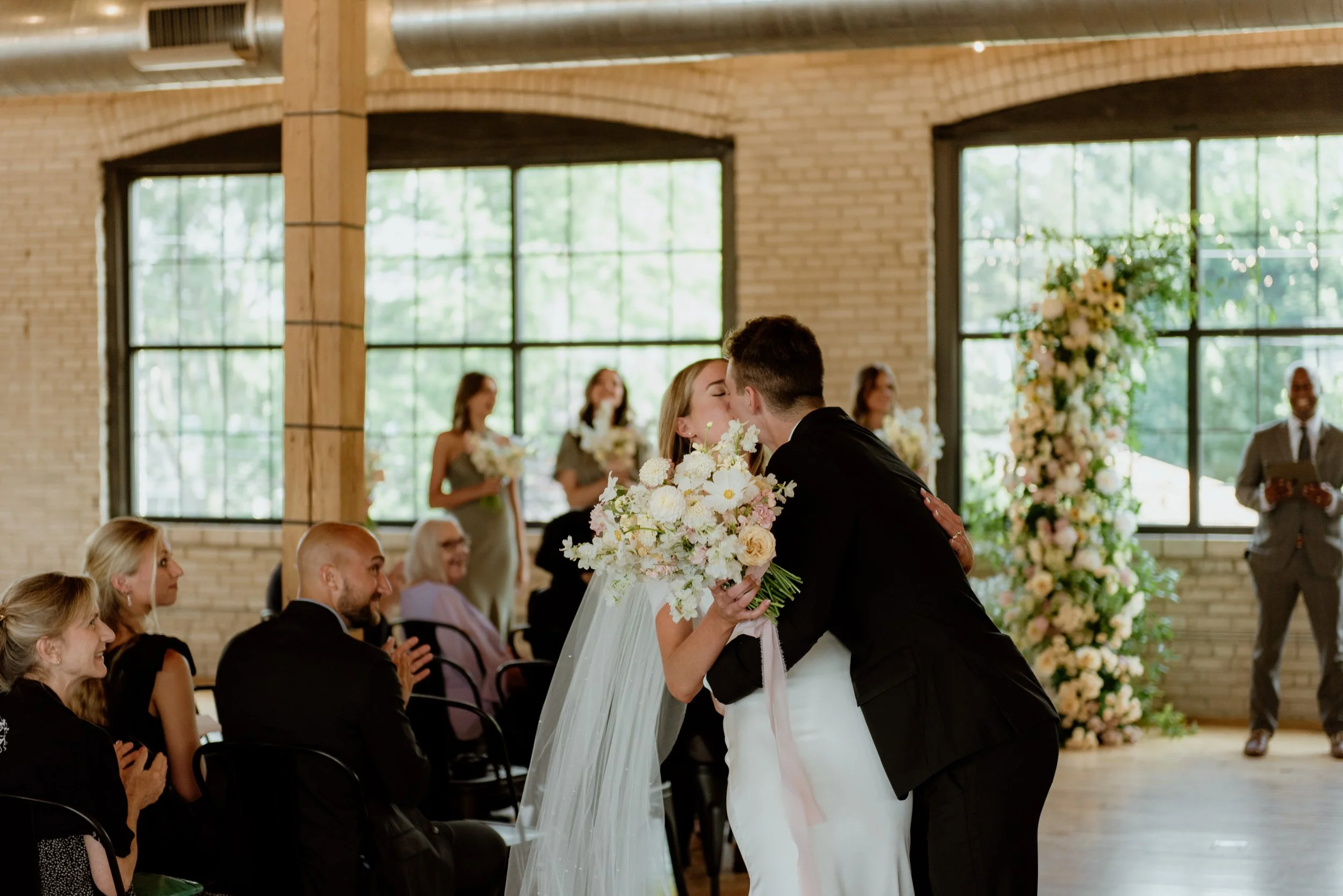 A bride and groom sharing a kiss during their wedding ceremony in a brightly lit indoor venue with large windows and exposed brick walls.