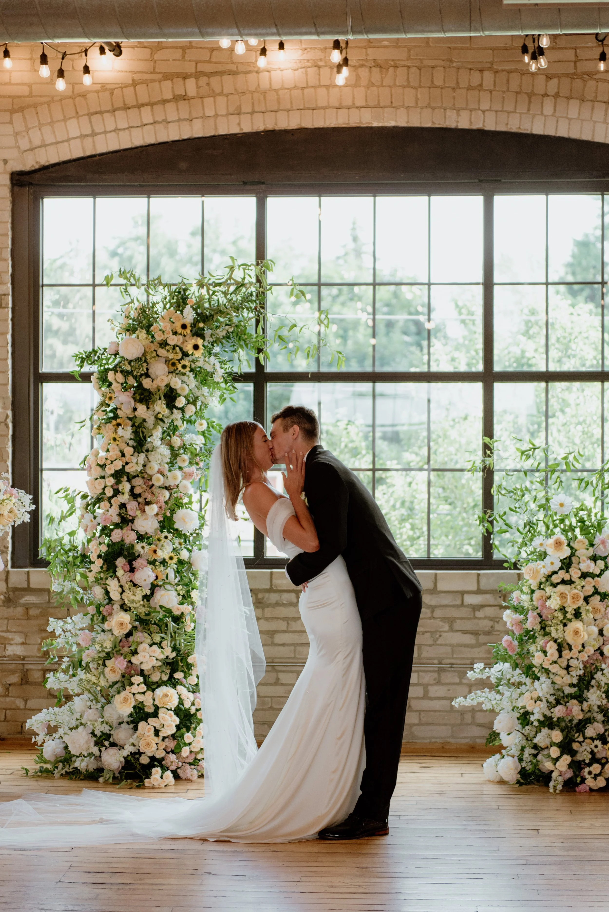 A bride and groom share a kiss during their wedding ceremony in a room with large windows and exposed brick walls, decorated with tall floral arrangements of white, pale pink, and green flowers.