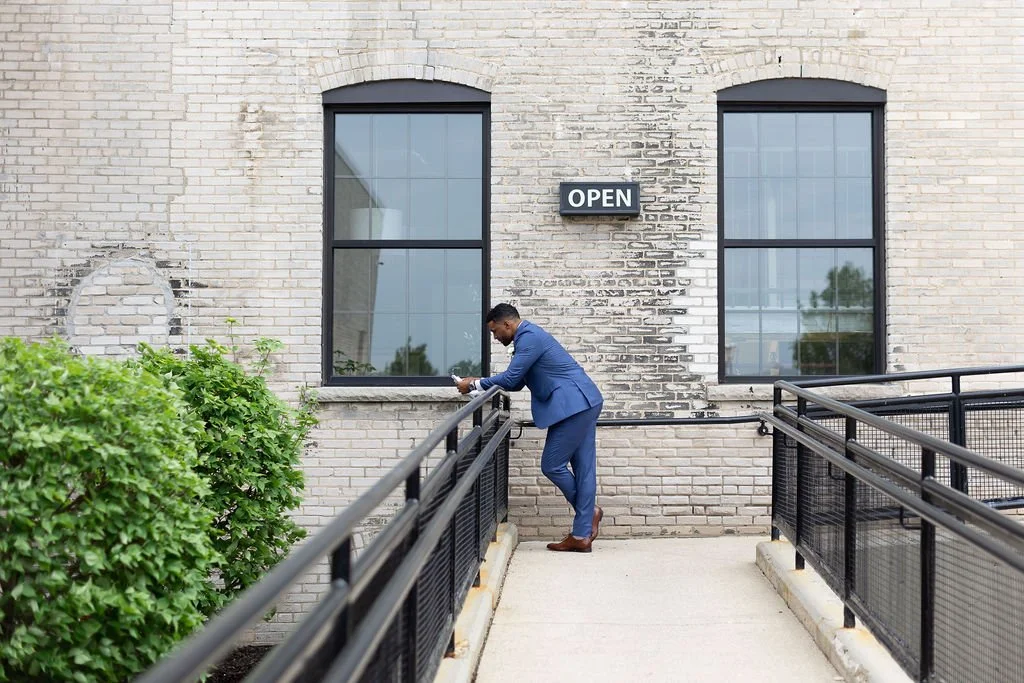 A man in a blue suit leaning on a black metal railing outside a brick building with large windows and an 'OPEN' sign.