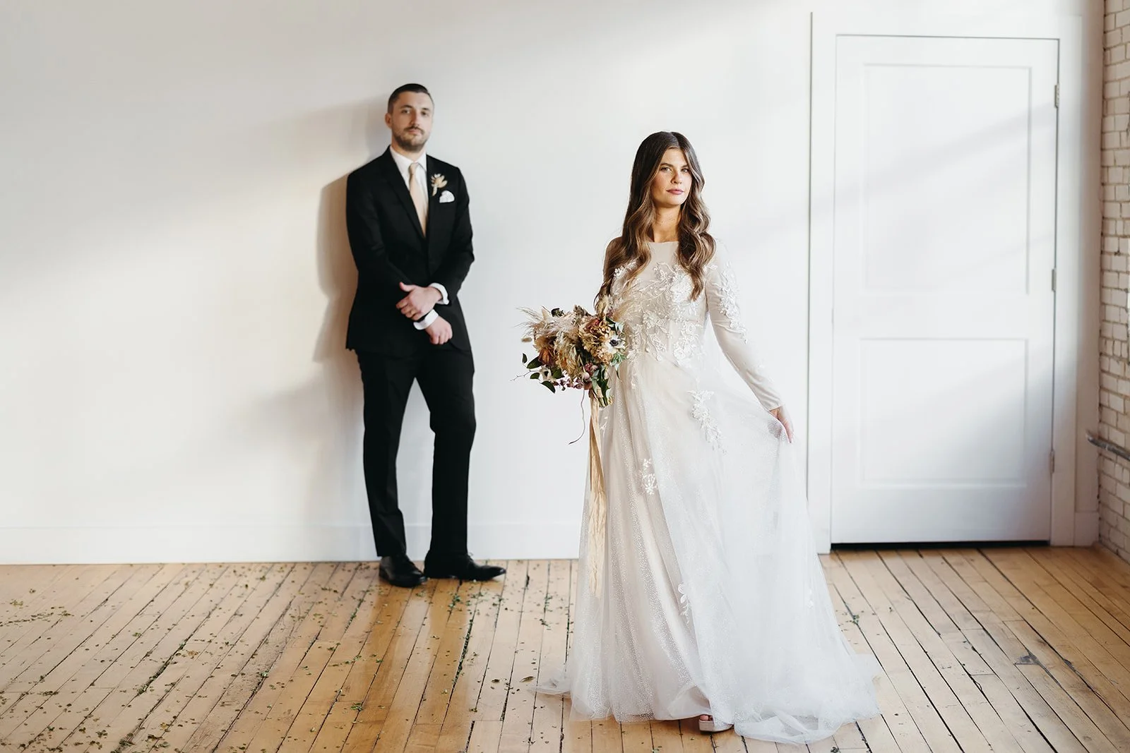 A bride in a white lace wedding dress holding a bouquet stands in foreground, while a groom in a black suit with a white tie and boutonnier leans against a light-colored wall in the background. The scene is in a minimalist room with a wooden floor an