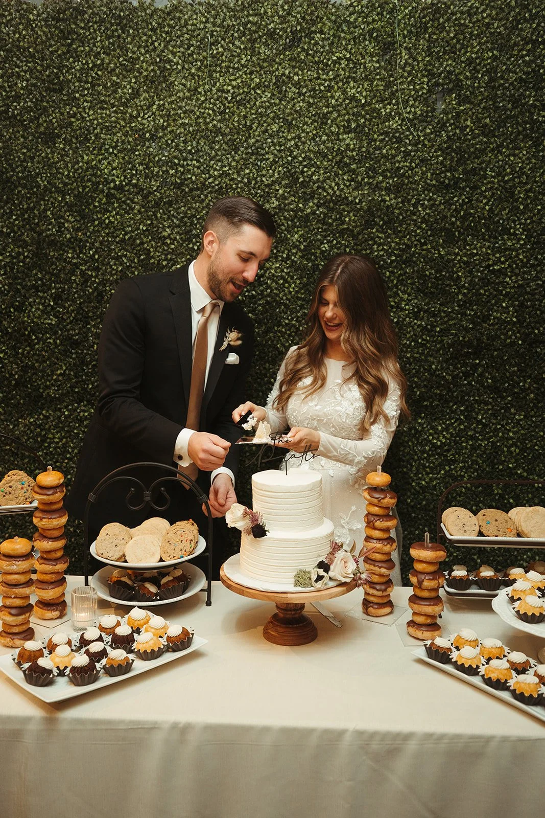 A bride and groom cutting a wedding cake at their reception, with dessert tables featuring cupcakes, cookies, and stacked biscuits nearby.