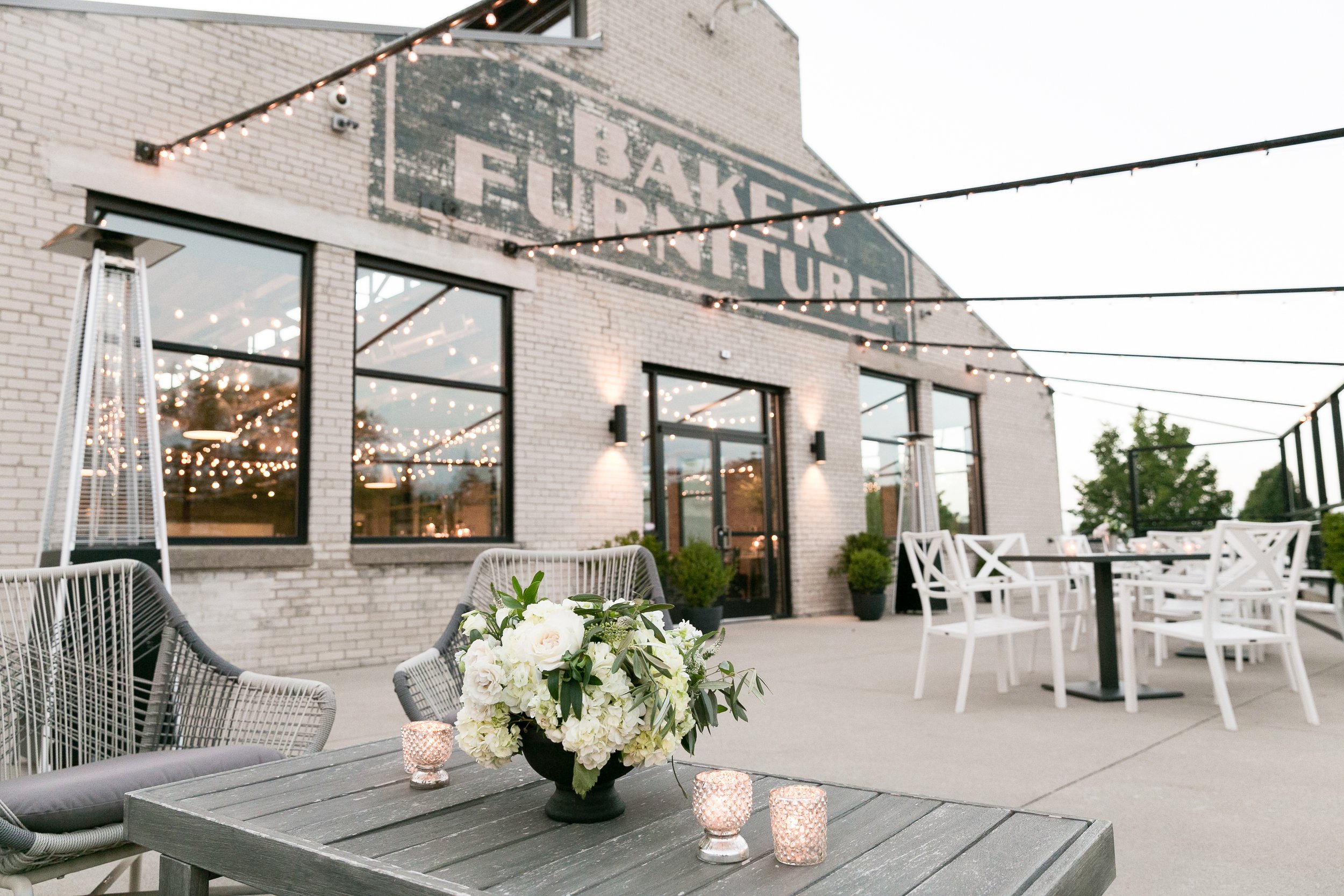Outdoor patio with white chairs and black tables, decorated with a flower arrangement and small candle holders. In the background, a brick building with large windows and string lights overhead. A sign on the building reads 'EAKERS LAURTURE'.