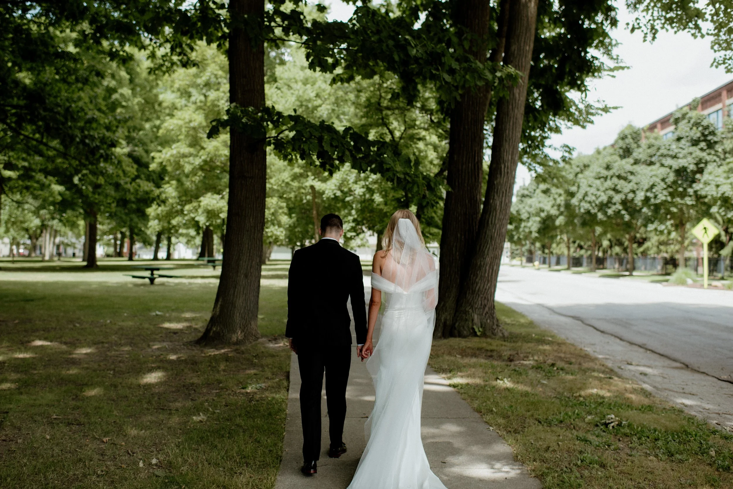 A bride and groom walking hand in hand through a park with tall green trees, the bride in a white wedding dress and veil, and the groom in a black suit.
