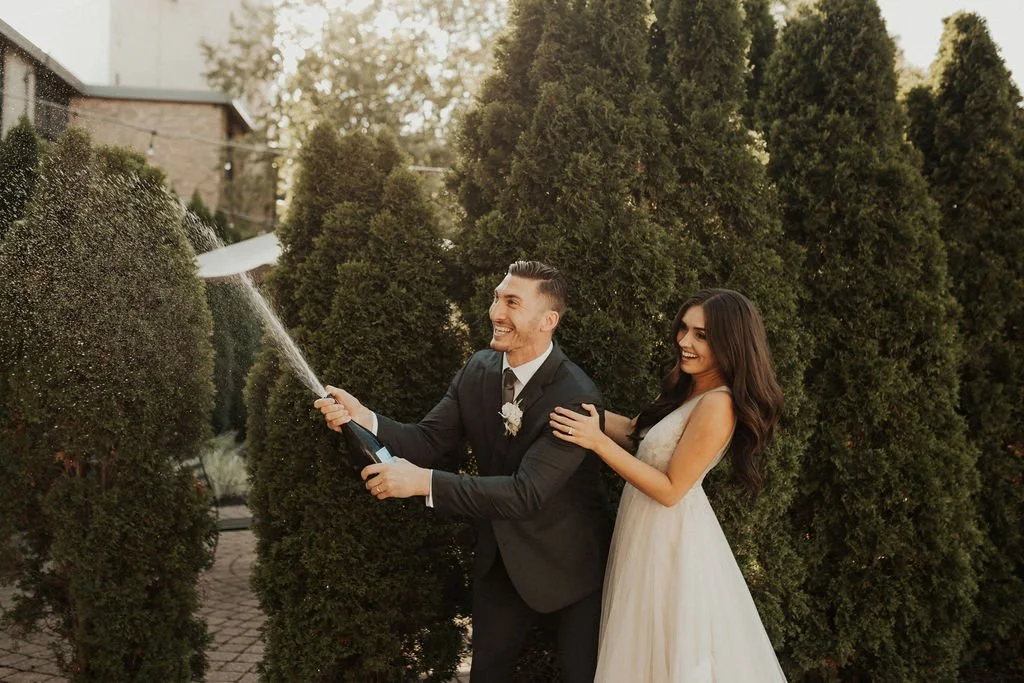 A newlywed couple celebrating outdoors on a sunny day, with the groom opening a champagne bottle and spraying it, while the bride stands behind him smiling and holding his shoulder. Tall green trees and a building are in the background.