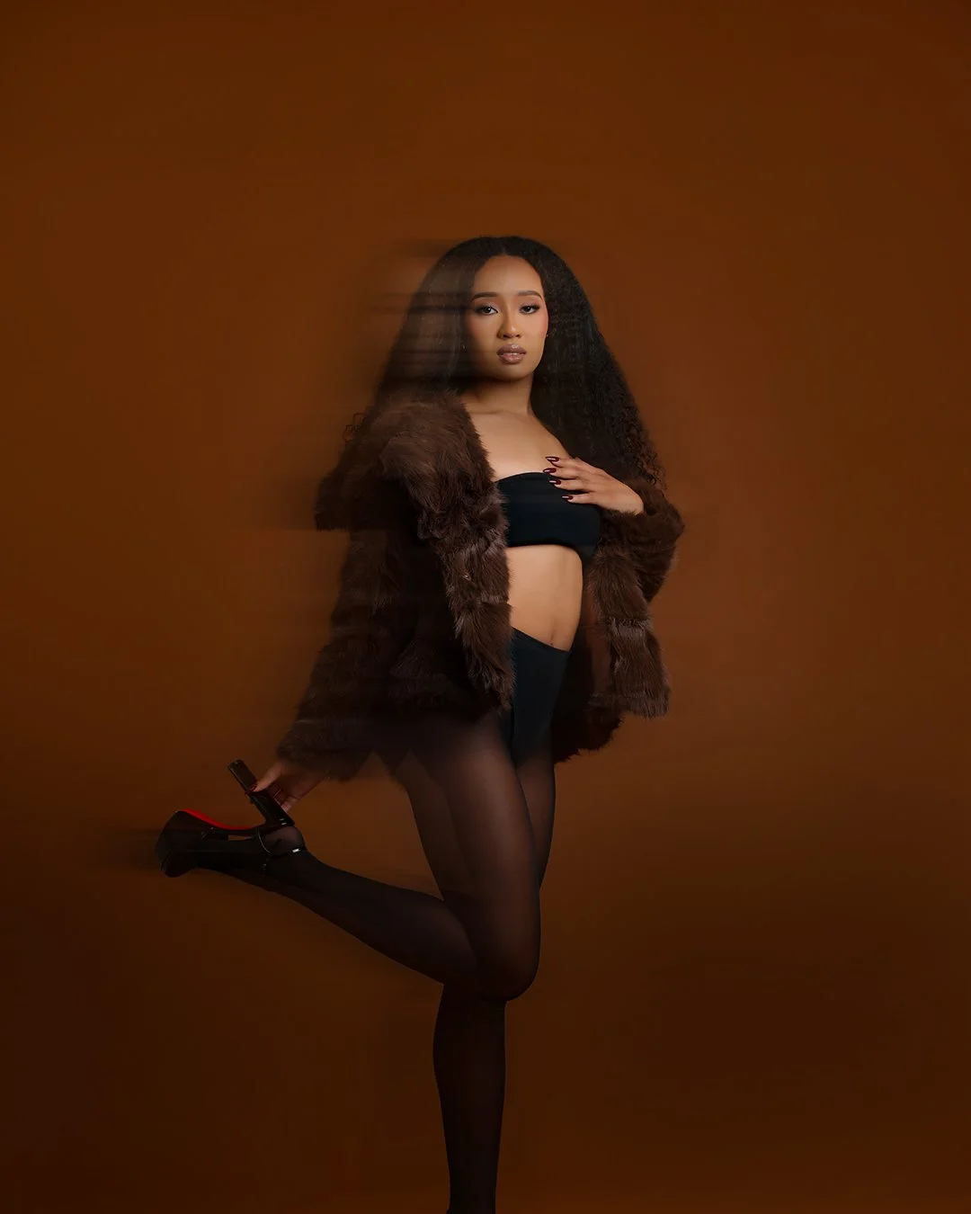 A woman with long curly hair wearing a black strapless top, sheer black tights, and black high heels, posing against a plain brown background.