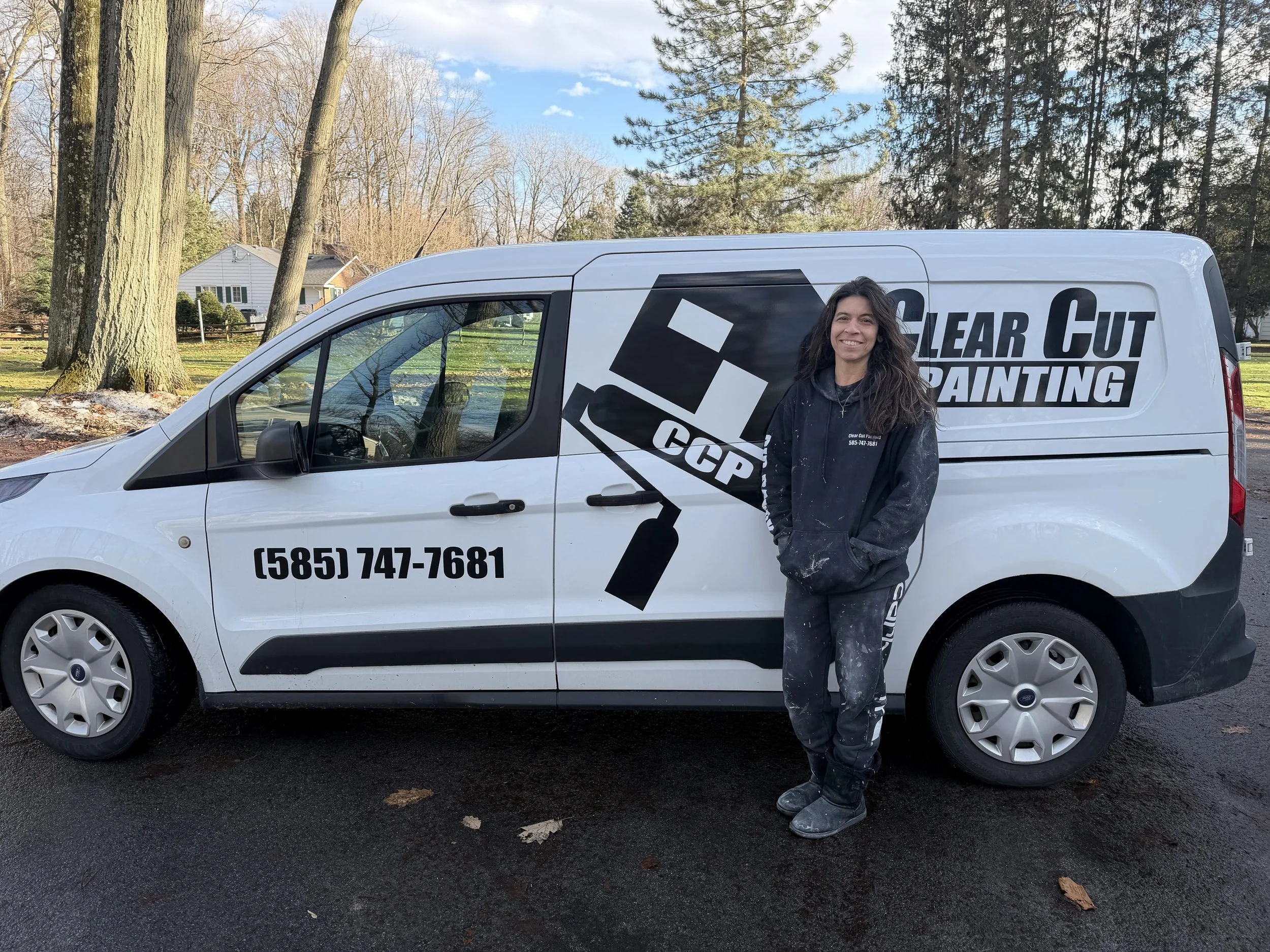 A woman standing next to a white van with a logo and text for Clear Cut Painting. She is wearing black work clothes with paint splatters, smiling with her hands in her pockets. The van displays a large paintbrush graphic, a phone number, and the company's name.