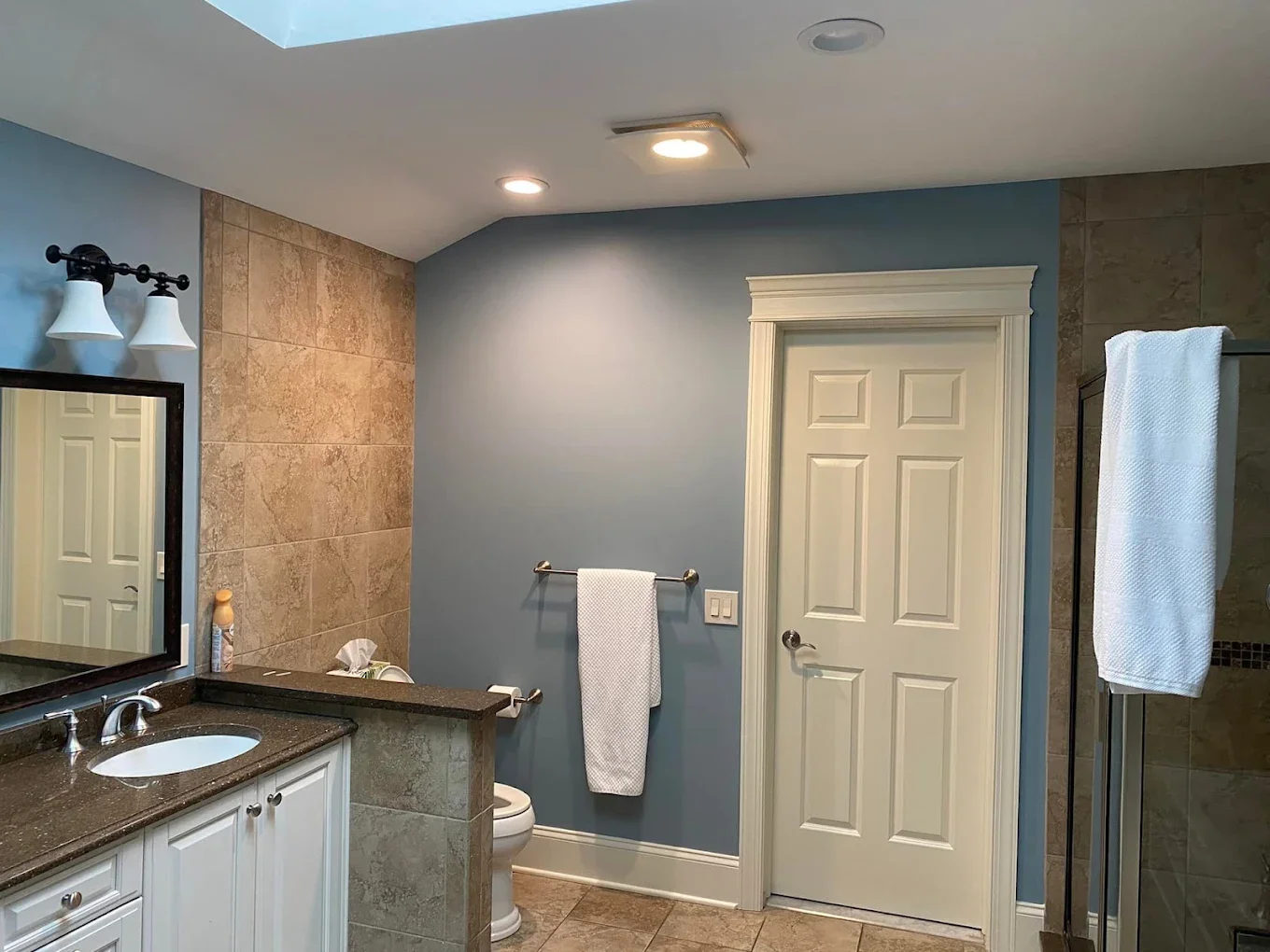 Bathroom with blue walls, a white door, a vanity with a black countertop, a mirror, a toilet, towel racks with towels, and a glass shower enclosure.
