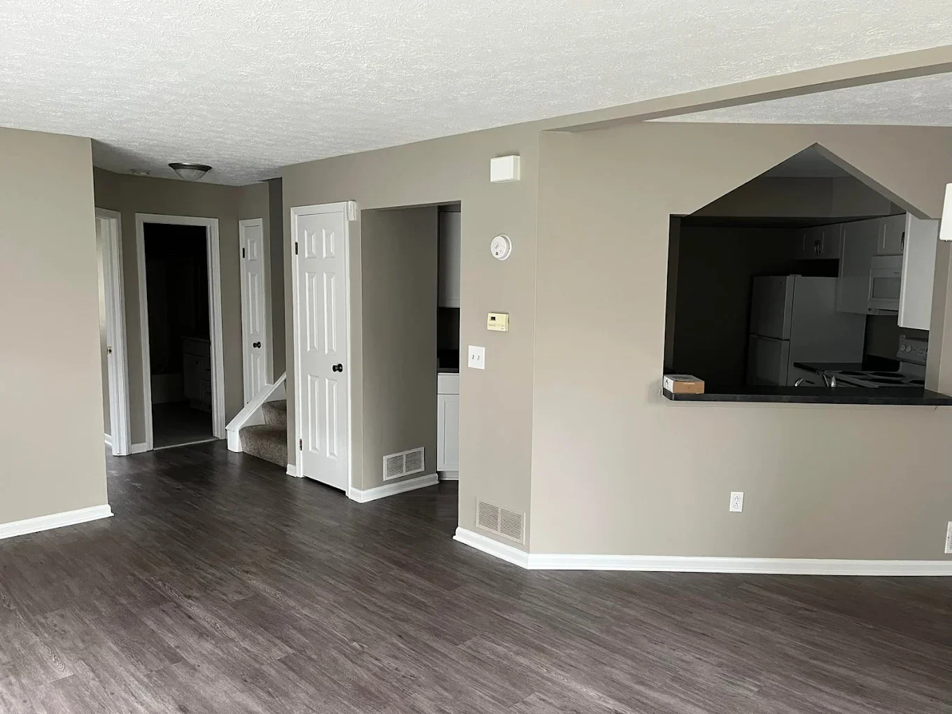 Empty living room with beige walls, dark hardwood flooring, white baseboards, and an open kitchen with a pass-through window.