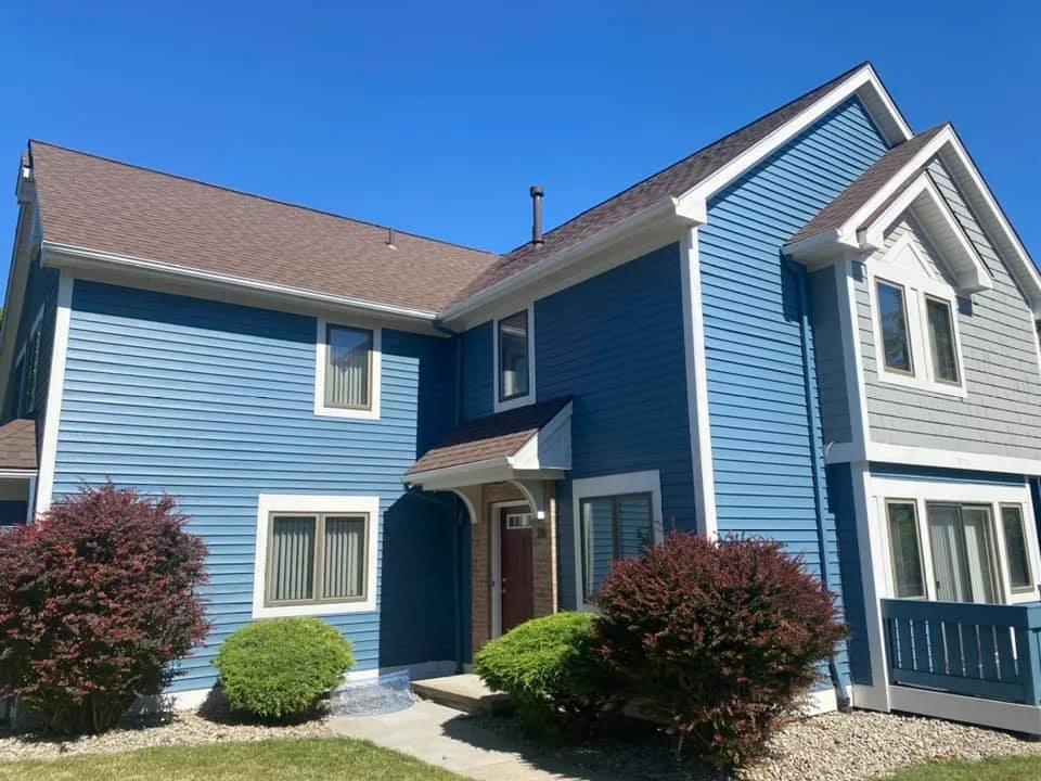 Blue two-story house with beige roof, white window trims, and landscaped front yard with shrubs and bushes, under a clear blue sky.
