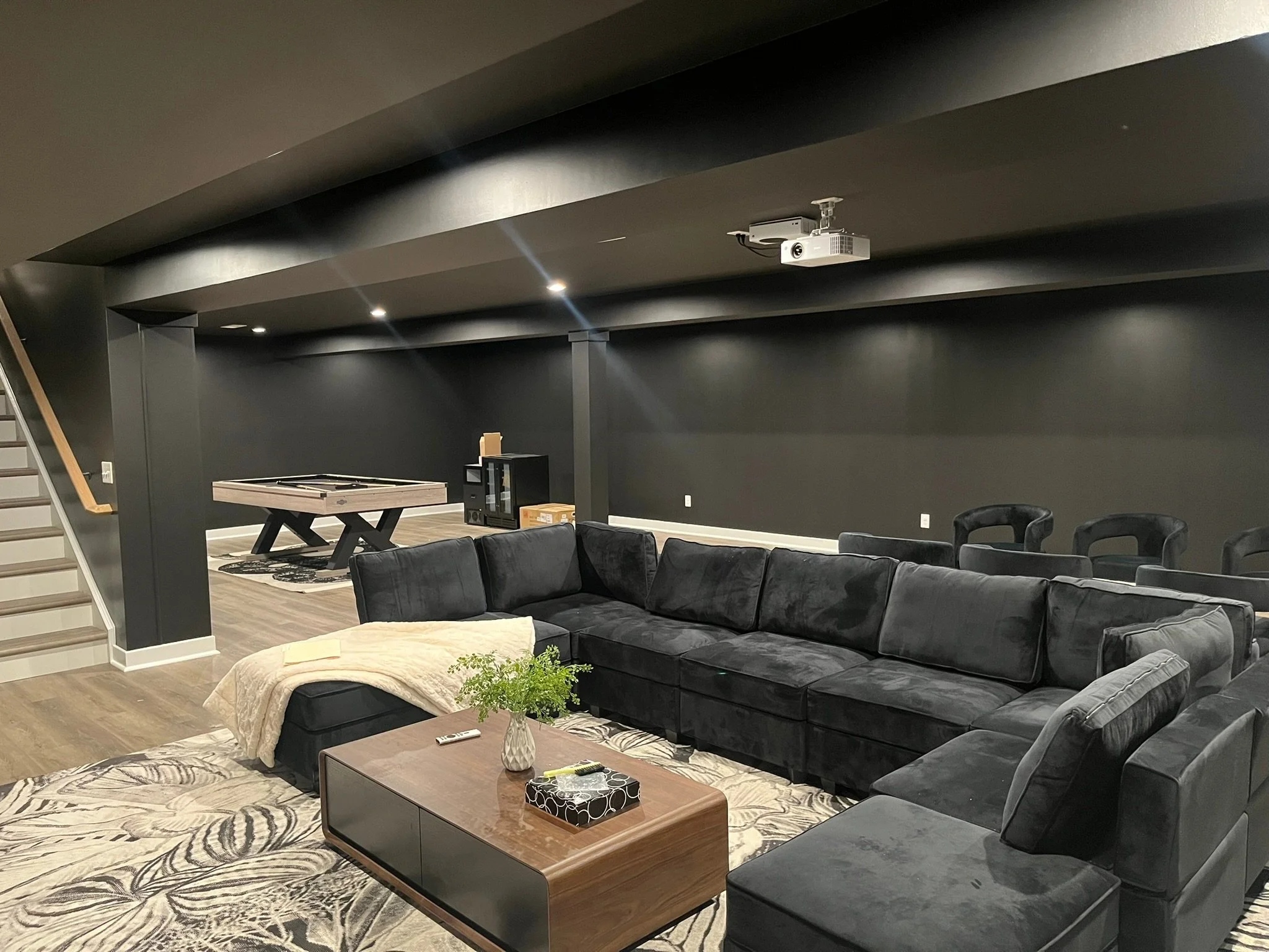 Living room with black walls, black sectional sofa, coffee table, patterned rug, and a game table in the background. Ceiling-mounted projector and chairs along the wall.